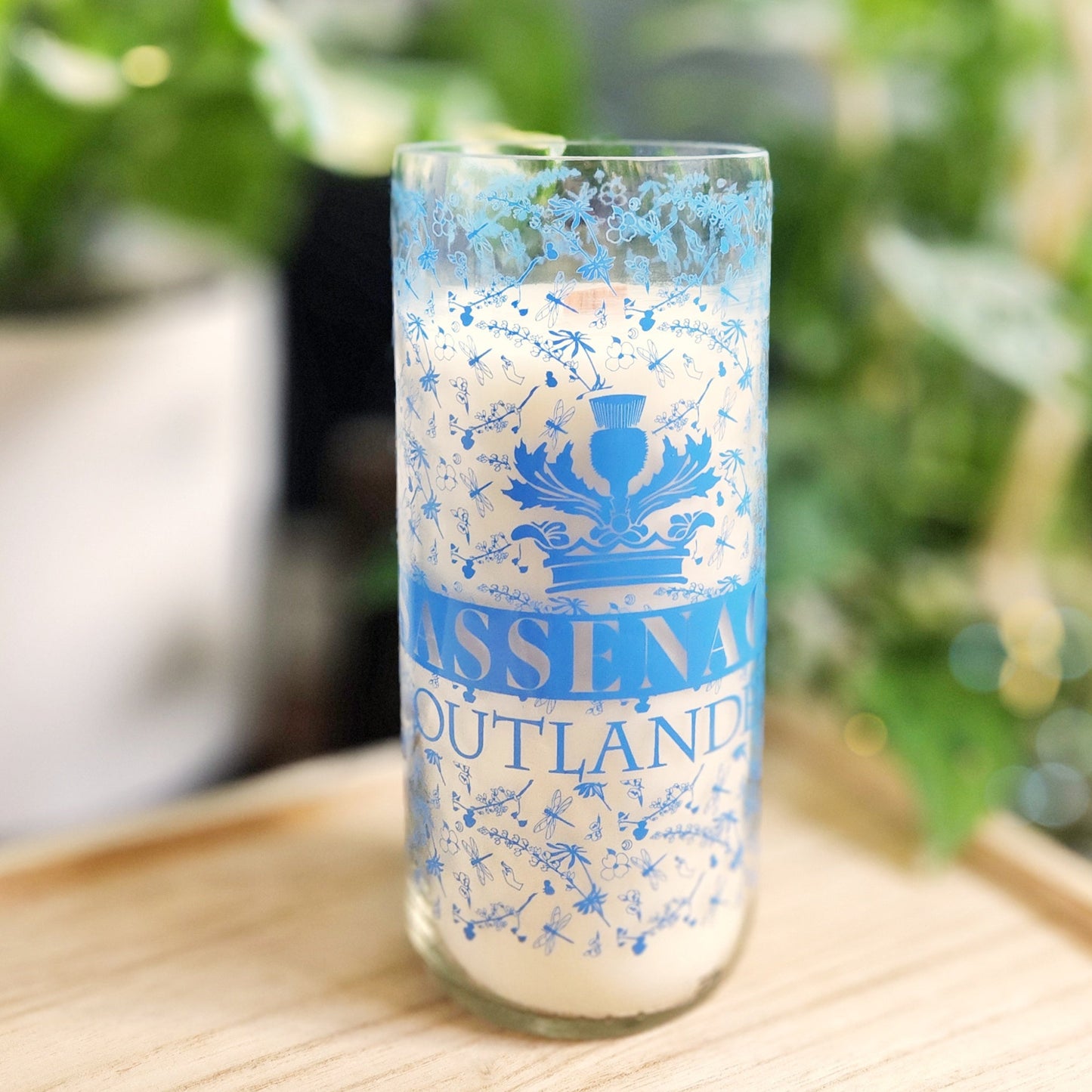 A hand-poured candle made from coconut-soy wax, housed in a recycled green wine bottle with a smooth, shiny rim. The bottle features a delicate blue floral design and the text "Sassenach Outlander," adding an elegant and vintage touch. The candle is set on a light wooden tray, surrounded by a soft-focus background of vibrant green plants, creating a serene and eco-friendly aesthetic. Perfect for showcasing sustainable, upcycled craftsmanship.