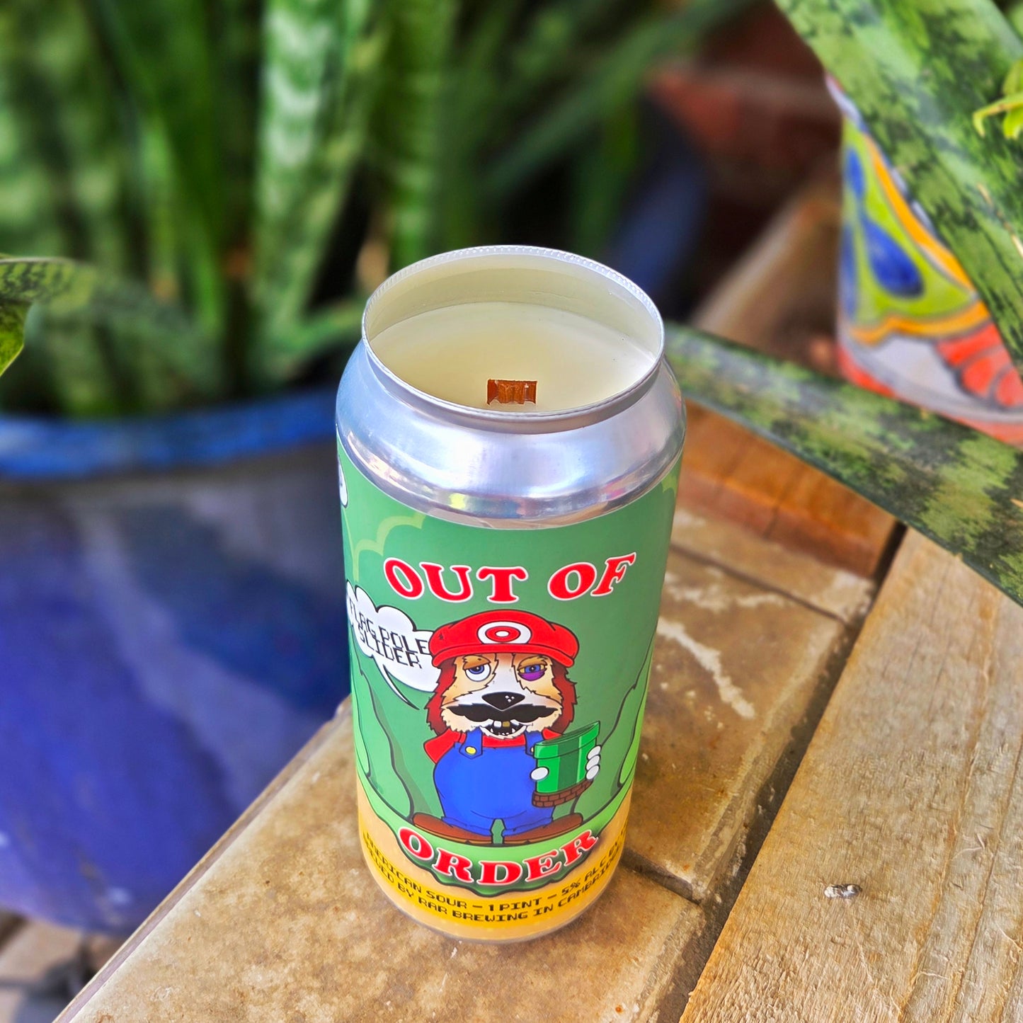 Angled shot of a handcrafted beer can candle in a reused RAR Brewing can with a “Flag Pole Slider” label featuring a dog dressed like a classic video game plumber. The coconut soy wax and wooden wick combo ensures a clean, cozy burn.
