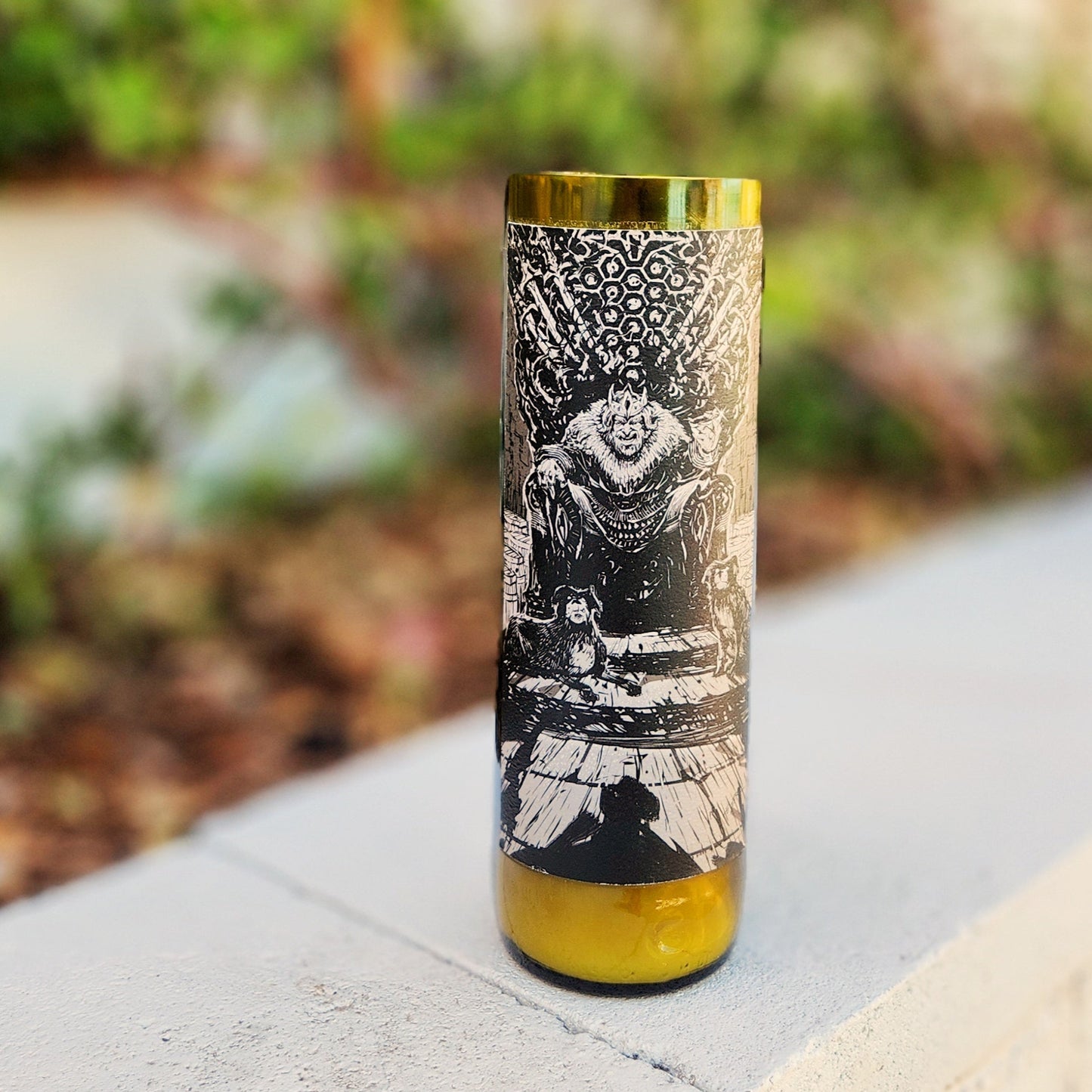A recycled glass candle crafted from a green wine bottle, featuring an intricate black-and-white label with an imposing illustration of a king-like figure seated on a throne adorned with ornate details. The label's dark and moody design adds a regal and mysterious vibe. The candle is placed outdoors on a light concrete surface, with blurred greenery in the background, enhancing its bold and striking appearance.