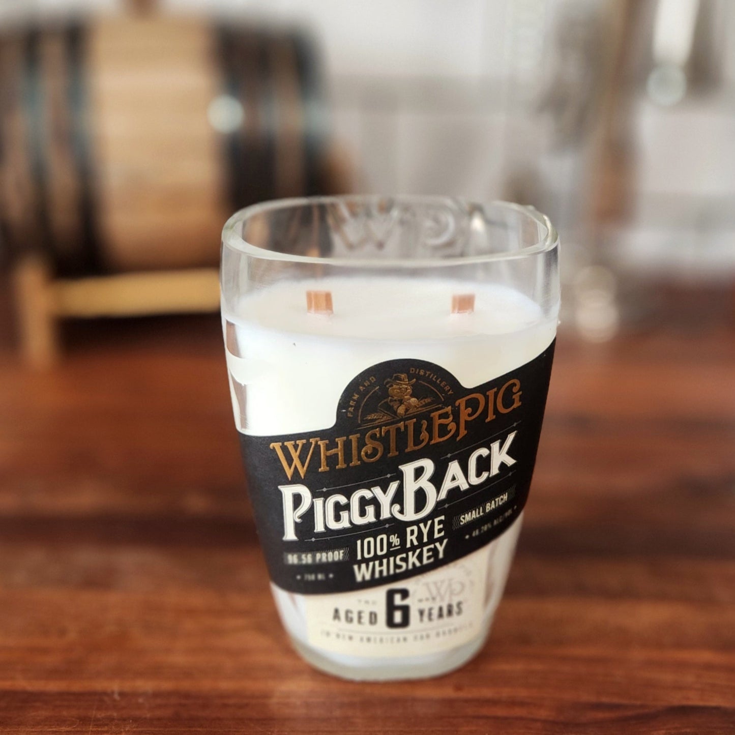 A hand-poured coconut-soy candle in a recycled WhistlePig PiggyBack whiskey bottle. Features double wooden wicks and the original black label. Displayed on a wooden surface with a blurred barrel backdrop, combining rustic charm and sustainability.