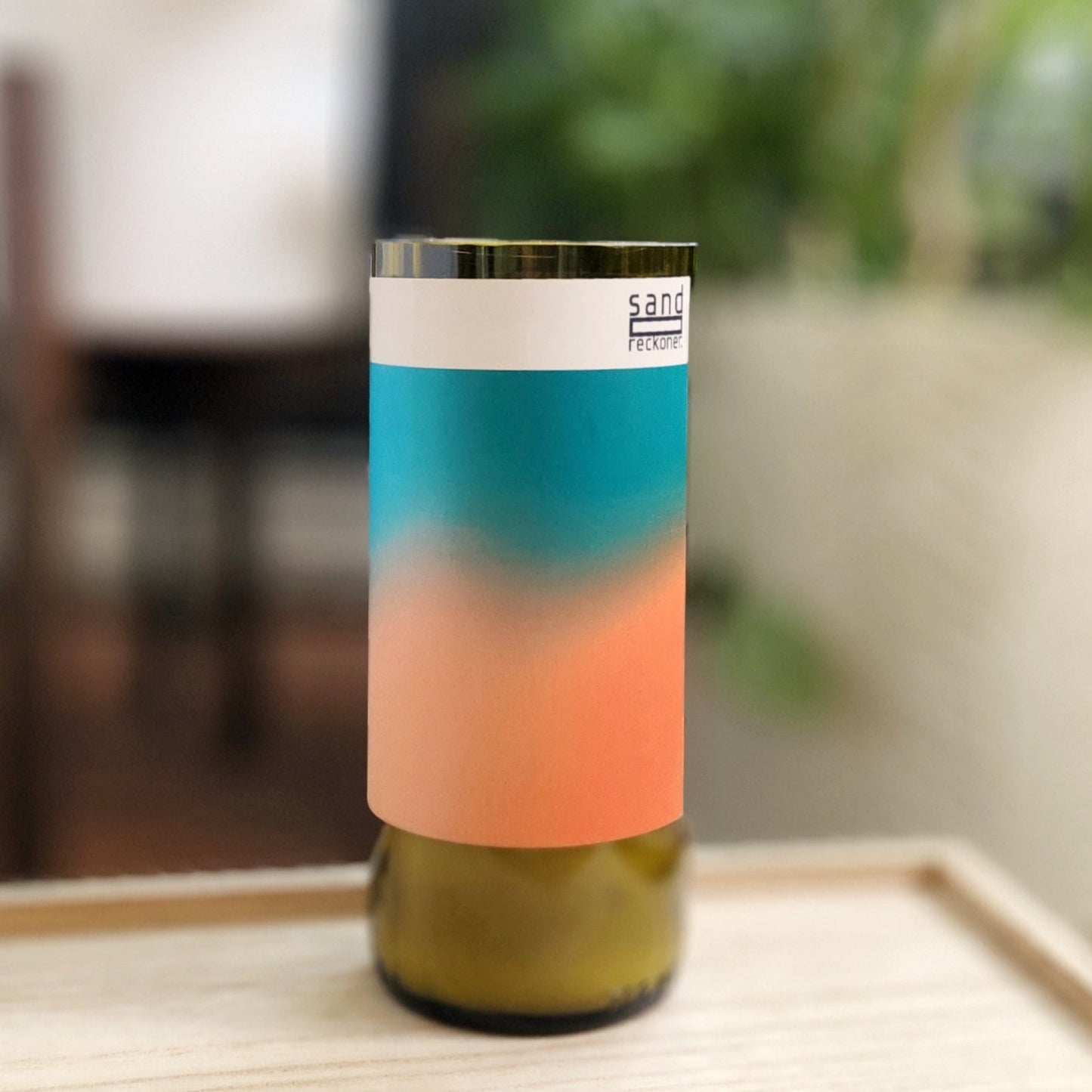A hand-poured candle crafted from coconut-soy wax, housed in a recycled green wine bottle with a smooth, polished rim. The label features a minimalist gradient design in soft orange and teal hues with the text "sand reckoner" at the top, adding a modern and artistic flair. The candle is displayed on a light wooden tray with a blurred background of green foliage and indoor decor, emphasizing its eco-friendly and stylish presentation. Perfect for sustainable and contemporary decor.