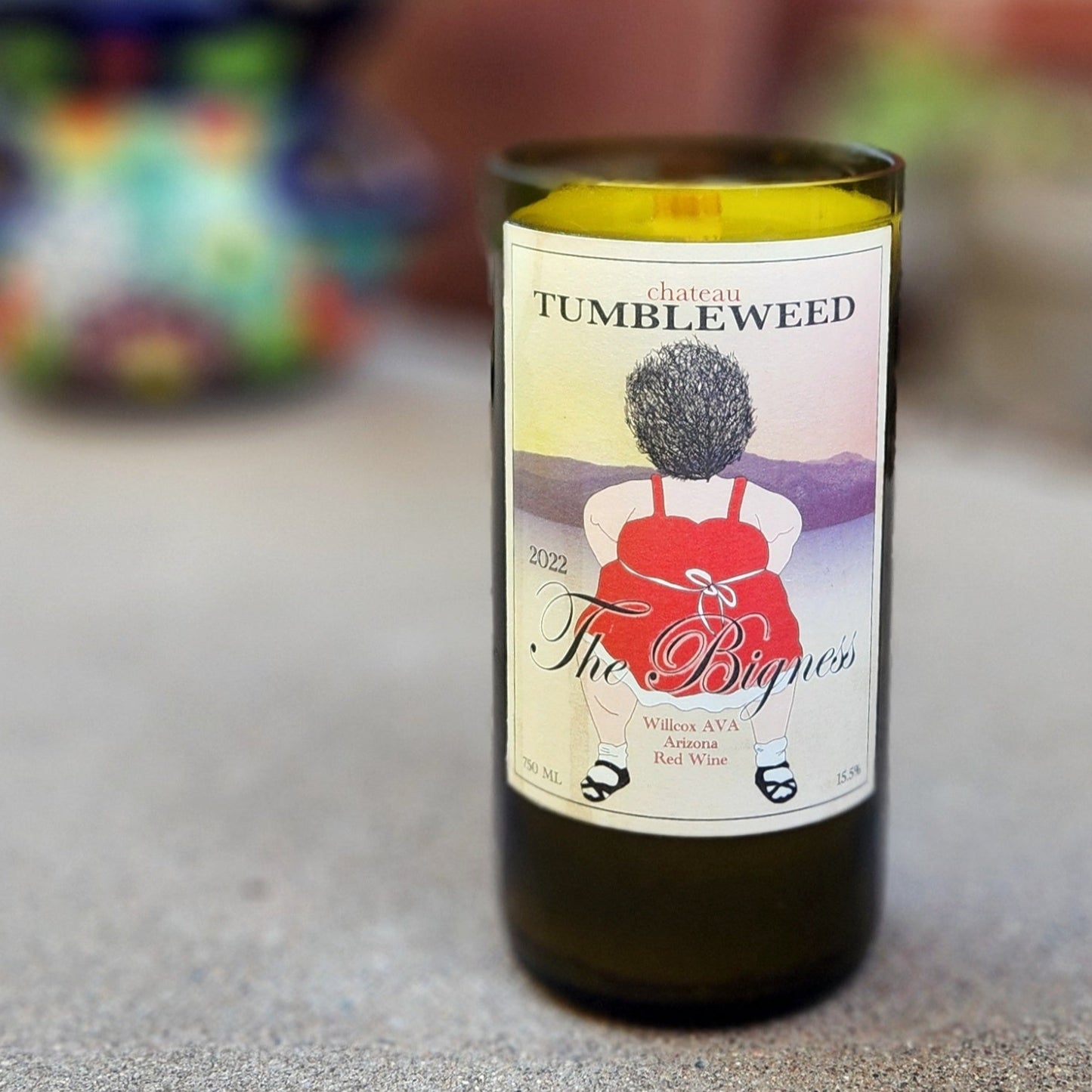 A handcrafted candle made from a recycled green wine bottle, featuring the original label from Chateau Tumbleweed's "The Bigness" 2022 Arizona Red Wine. The label showcases a whimsical illustration of a figure in a red dress with a tumbleweed for a head, set against a soft gradient of desert hues. The candle rests on a light concrete surface outdoors, with a blurred background of potted plants and a vibrant ceramic planter, evoking a colorful and artistic Southwestern charm.