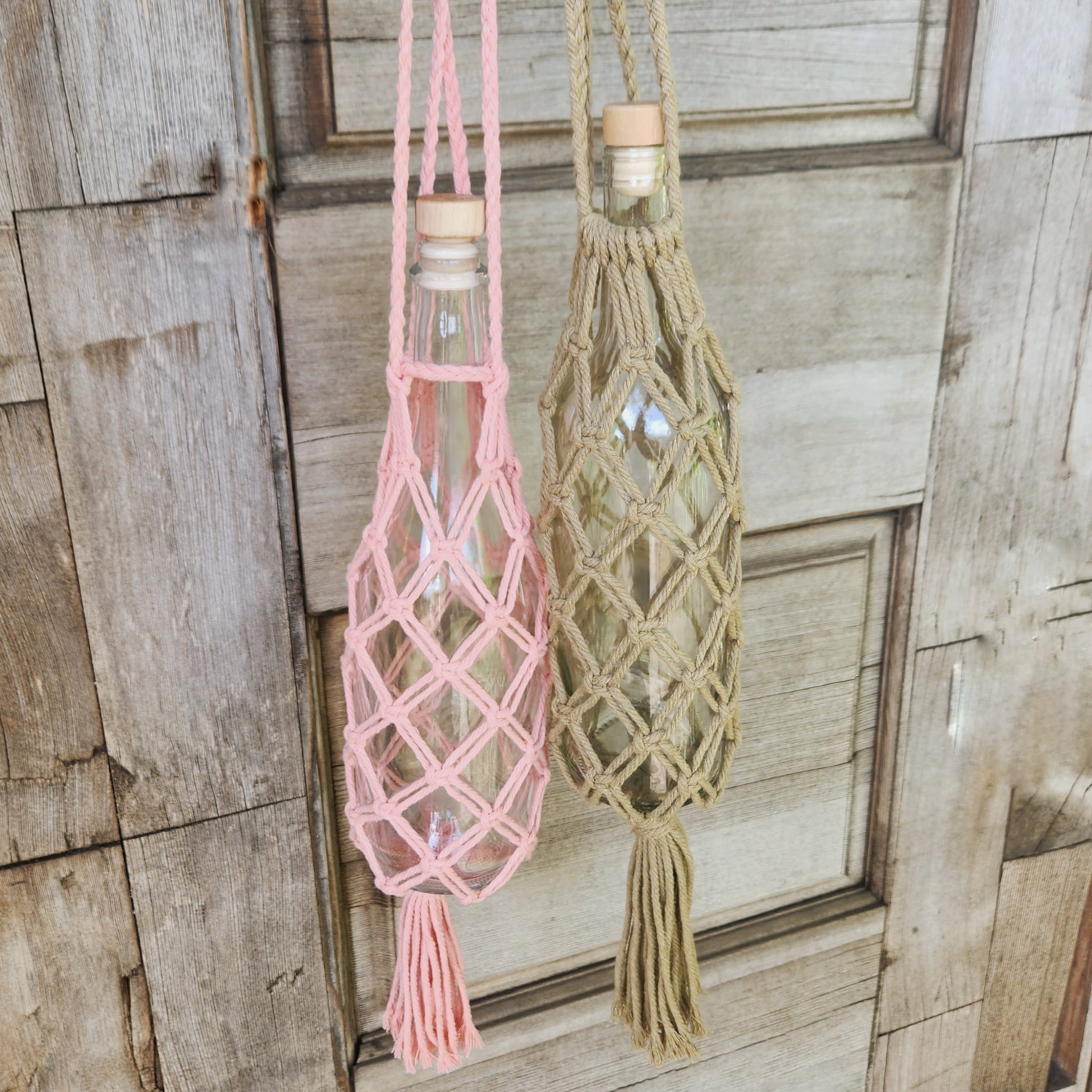 Close-up of two handmade macrame wine bottle tote bags hanging against a rustic wooden backdrop, showcasing intricate knot patterns in beige and pink colors