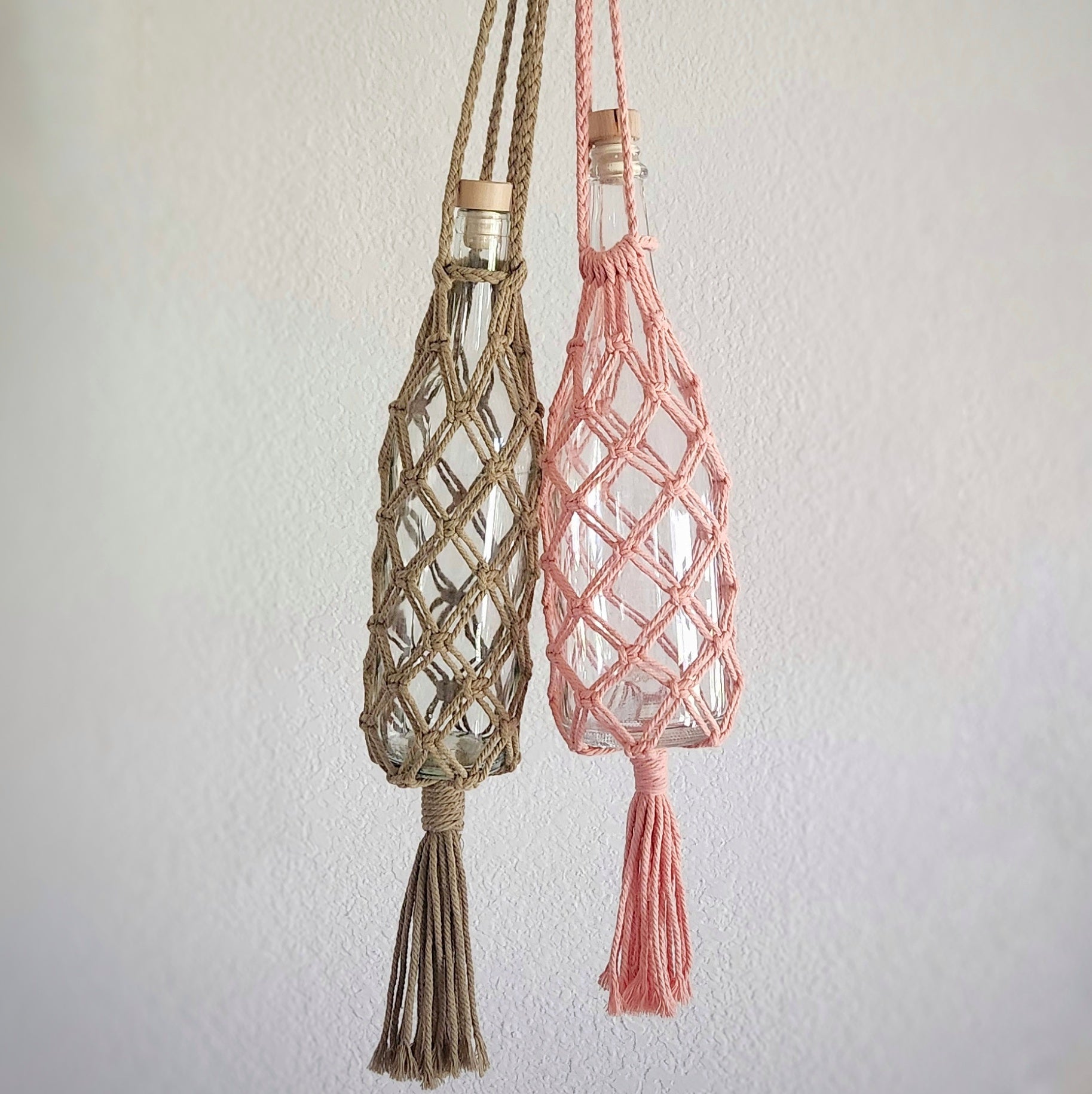 two macrame wine bottle tote bags hanging on a plain wall, featuring detailed knot designs in sage green and pale salmon.