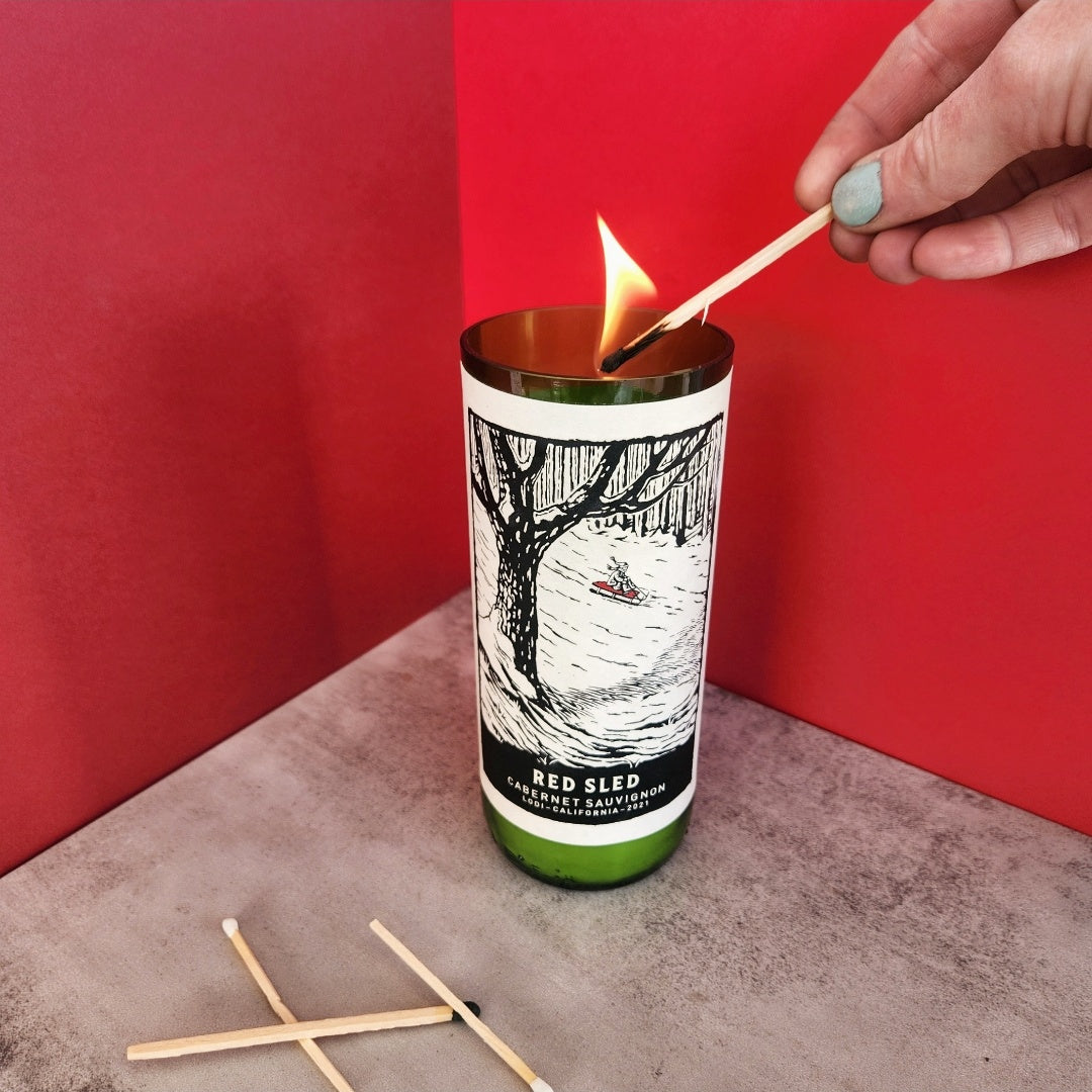 A Cactus+Vine Fir Tree scented candle in a recycled Red Sled wine bottle being lit with a match, showcasing the elegant design and eco-friendly materials.