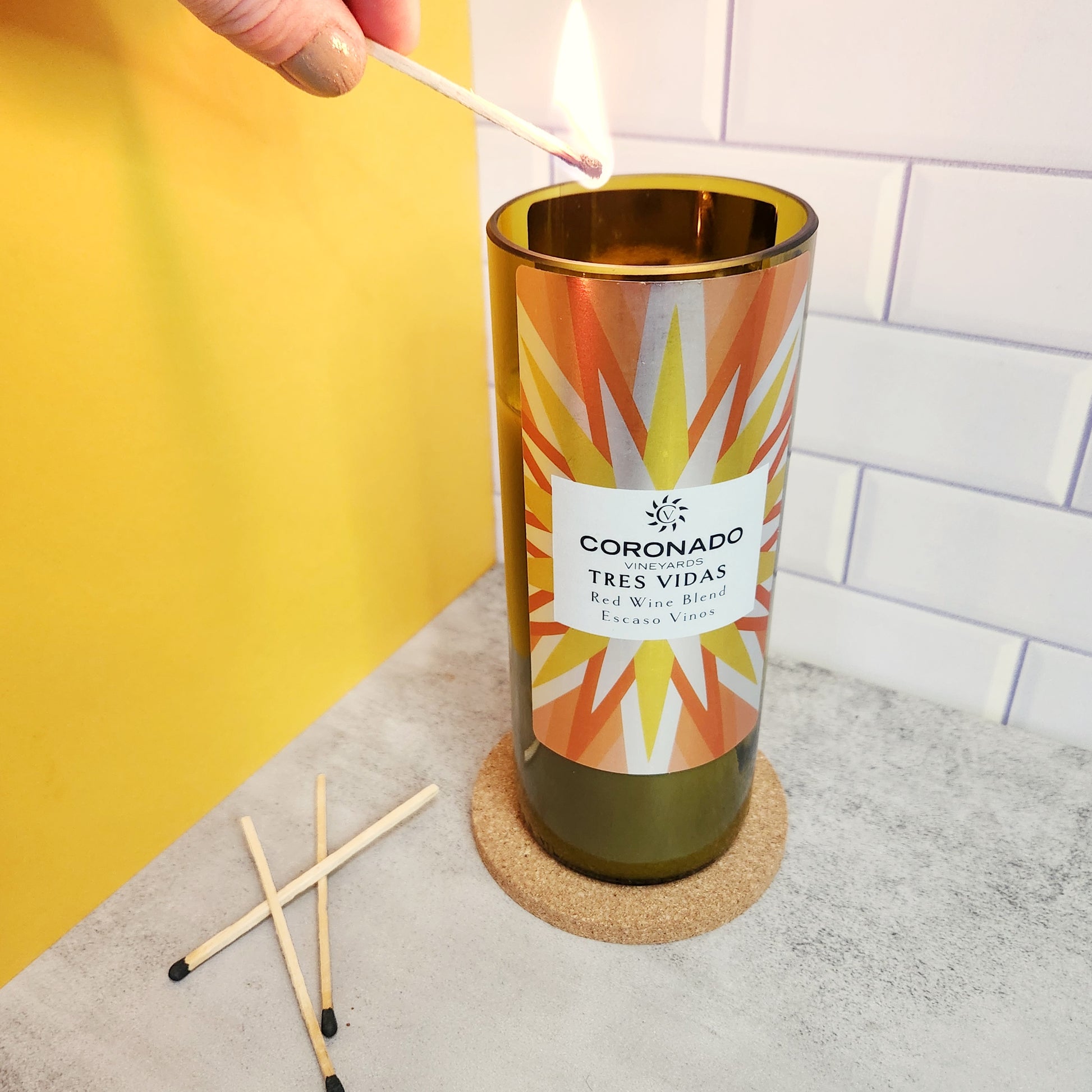 Hand lighting a wooden wick candle made from a recycled Coronado Vineyards Tres Vidas wine bottle, featuring a vibrant label design for sustainable home décor.