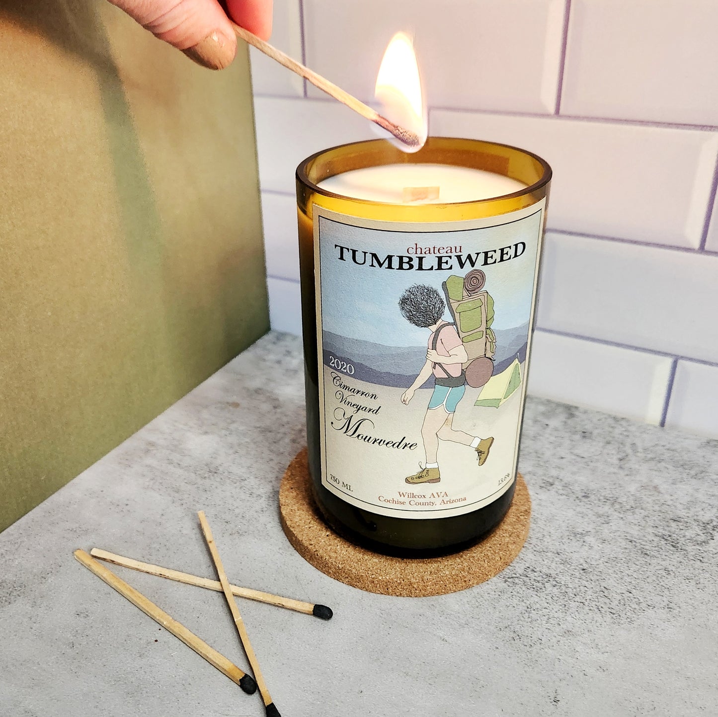 Hand lighting a Chateau Tumbleweed candle made from a recycled wine bottle with a wooden wick, showcasing a unique hiking-themed label design.