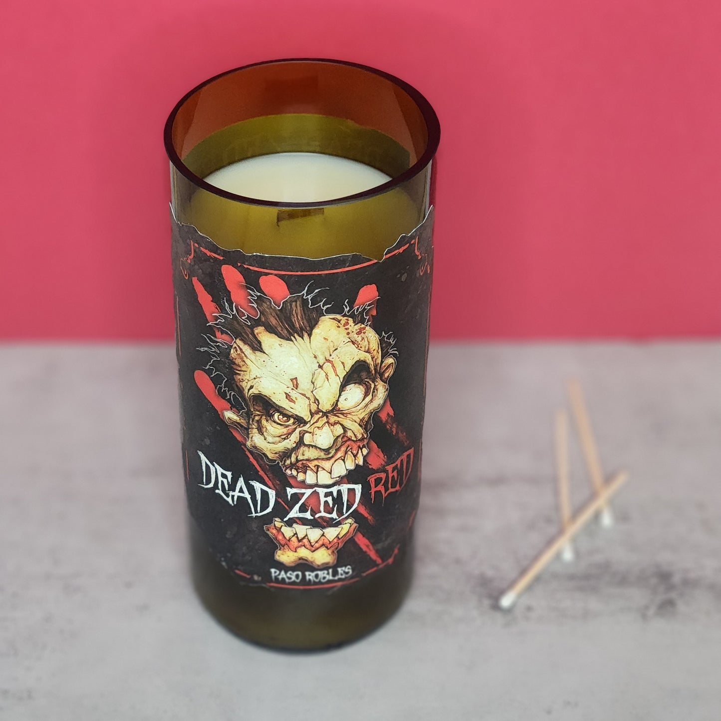 Dead Zed Red wine bottle candle with a wooden wick, set against a red background with matches, highlighting its recycled wine bottle design and sustainable qualities, ideal for Halloween decor.