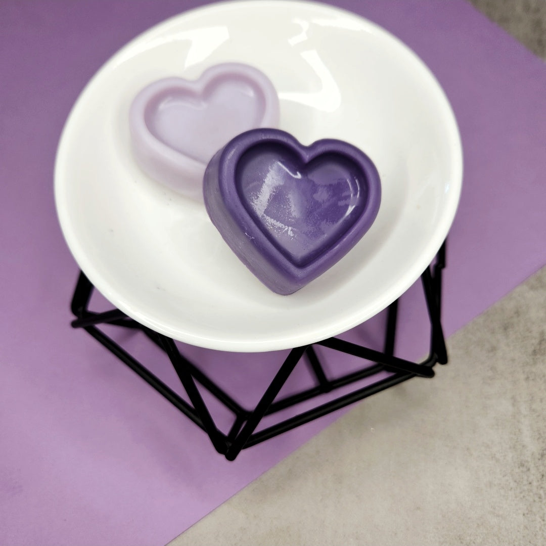 Two heart-shaped Lavender Library wax melts in different shades of purple displayed on a white wax melter with a black geometric stand, set against a purple background for a calming and stylish presentation