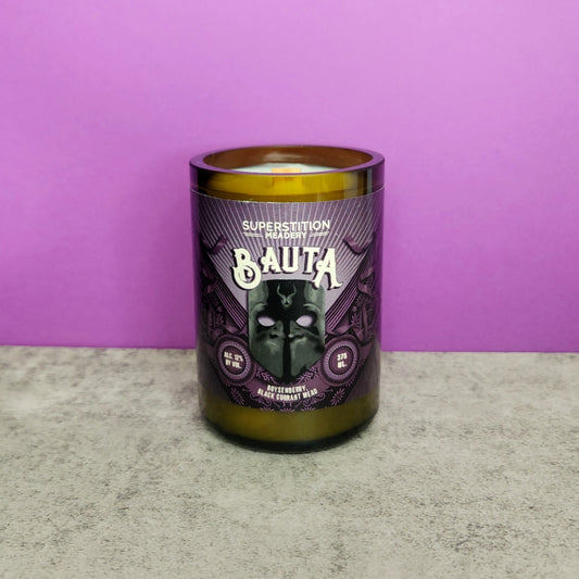 Recycled Arizona mead bottle candle hand-poured by Cactus Vine Candles featuring a Superstition Meadery Bauta label. The candle is set on a light gray surface against a purple background, highlighting its eco-friendly, coconut soy blend and bold label design. The label showcases a gothic-inspired mask with dark, rich colors, adding a mysterious and dramatic touch to the candle.