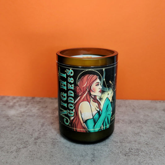 Recycled Arizona mead bottle candle hand-poured by Cactus Vine Candles featuring a vibrant Night Goddess label. The candle is set on a light gray surface against an orange background, highlighting its unique design and eco-friendly, coconut soy blend. The label showcases an illustrated woman with red hair, evoking a mystical and celestial theme.