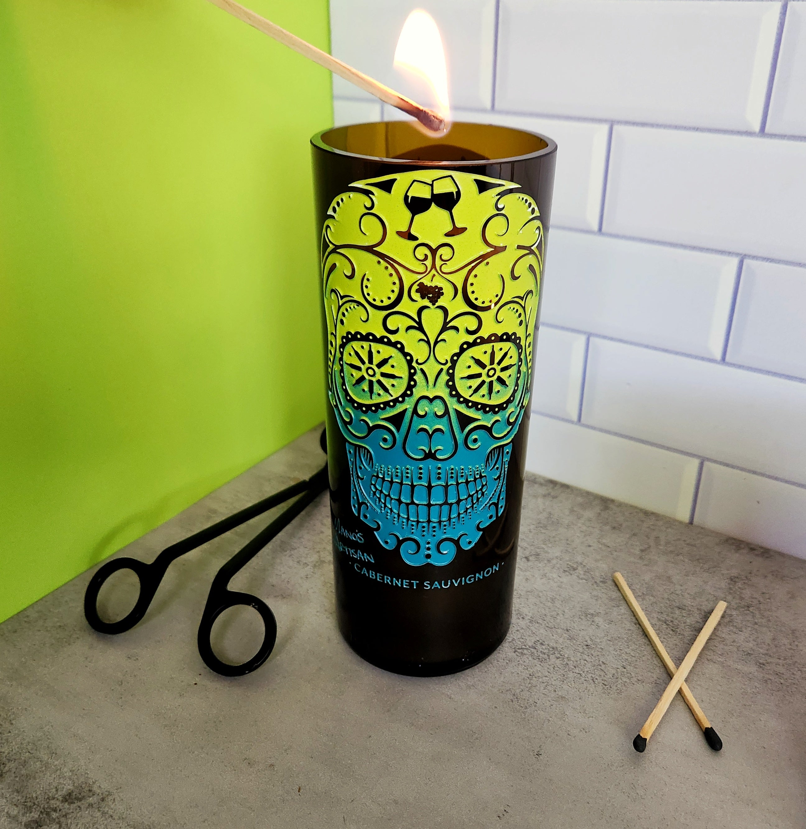 Colorful skull design wine bottle candle being lit with matches, showcasing handmade recycled wine bottle candles – Day of the Dead decor inspiration.