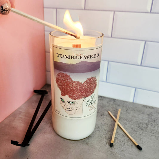 16oz Strawberry Wine scented candle being lit, housed in a recycled Chateau Tumbleweed wine bottle. The candle features a unique label design with artistic illustrations, adding a touch of whimsy to the modern decor. Two unlit matches and a black candle wick trimmer are placed beside the candle.