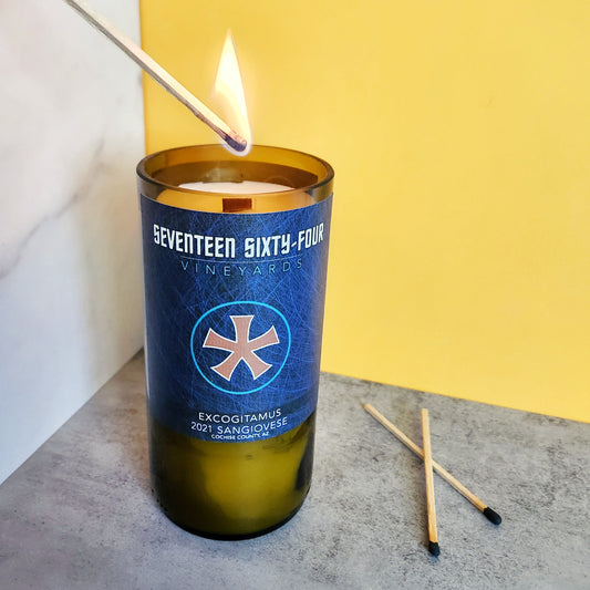 16oz House Red scented wine bottle candle being lit, housed in a recycled 1764 Vineyards wine bottle. The label features a blue patterened design with the 1764 Vineyards logo, adding an elegant touch to this Arizona wine bottle candle. Perfect for adding a rich, fruity aroma to your home decor.