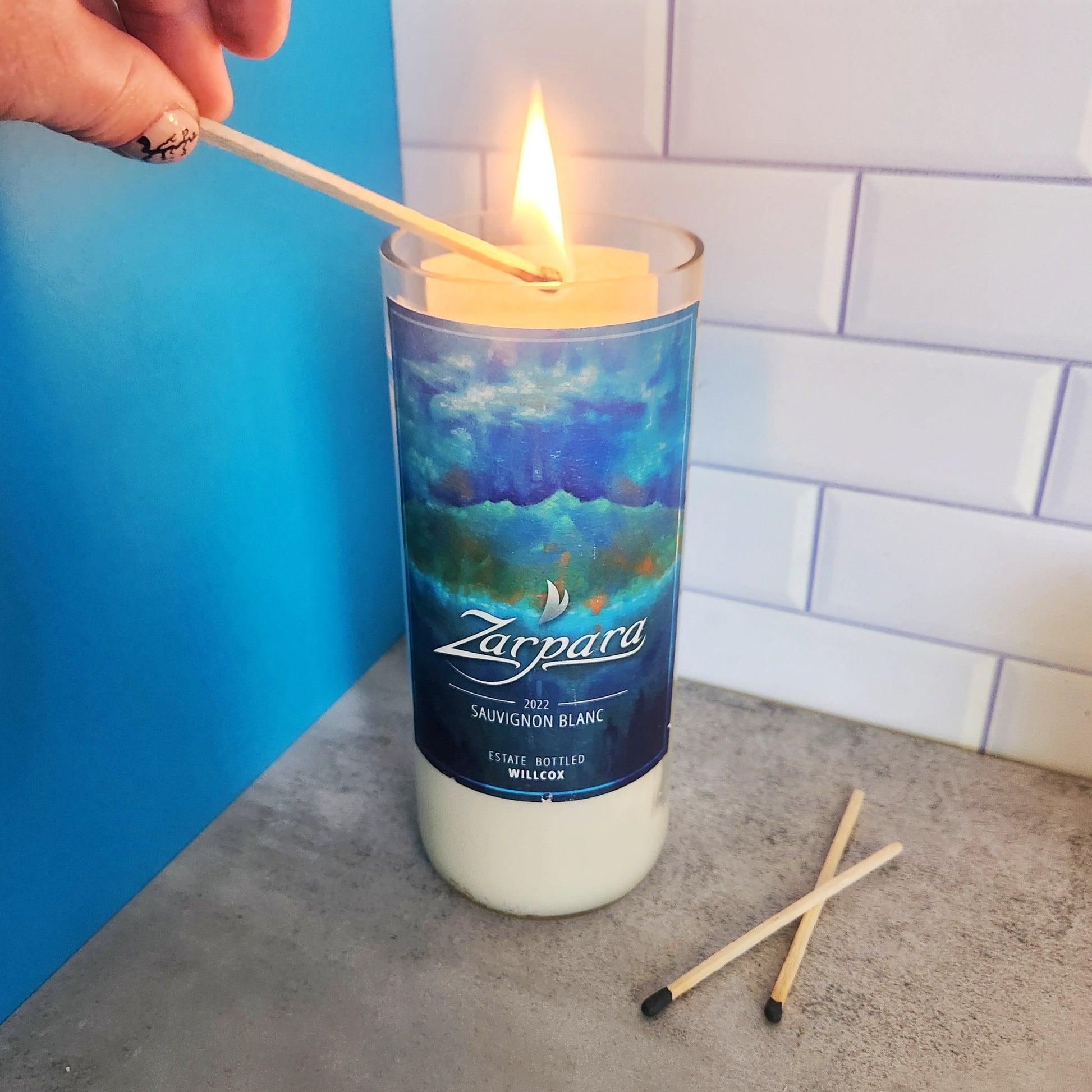 Hand lighting a Cactus Vine Candle in a recycled Arizona wine bottle with a Zarpára Sauvignon Blanc label. The candle is positioned on a light gray surface with two matches nearby, and the background features blue and white tiles.