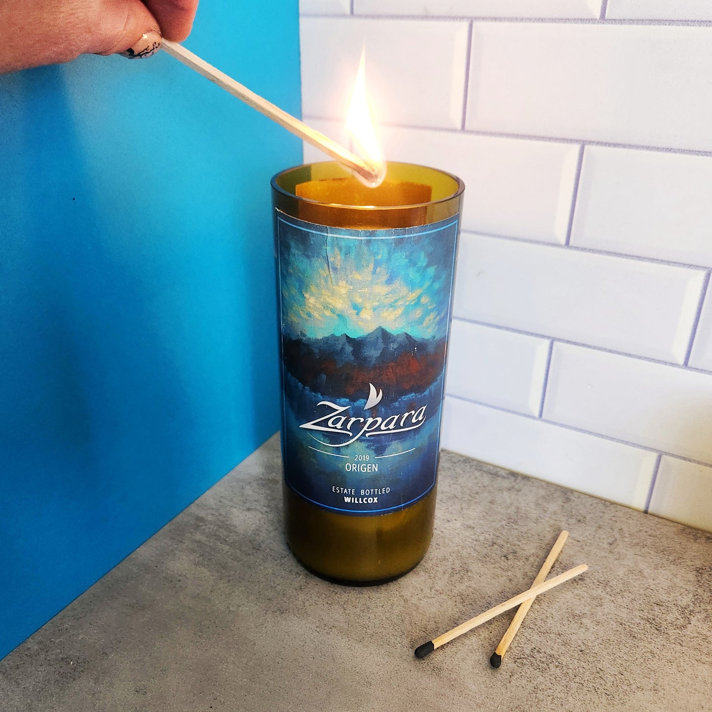 Hand lighting a wine bottle candle in a recycled Arizona wine bottle with a Zarpára Origen label. The candle is set on a light gray surface with two matches nearby, and the background features blue and white tile elements