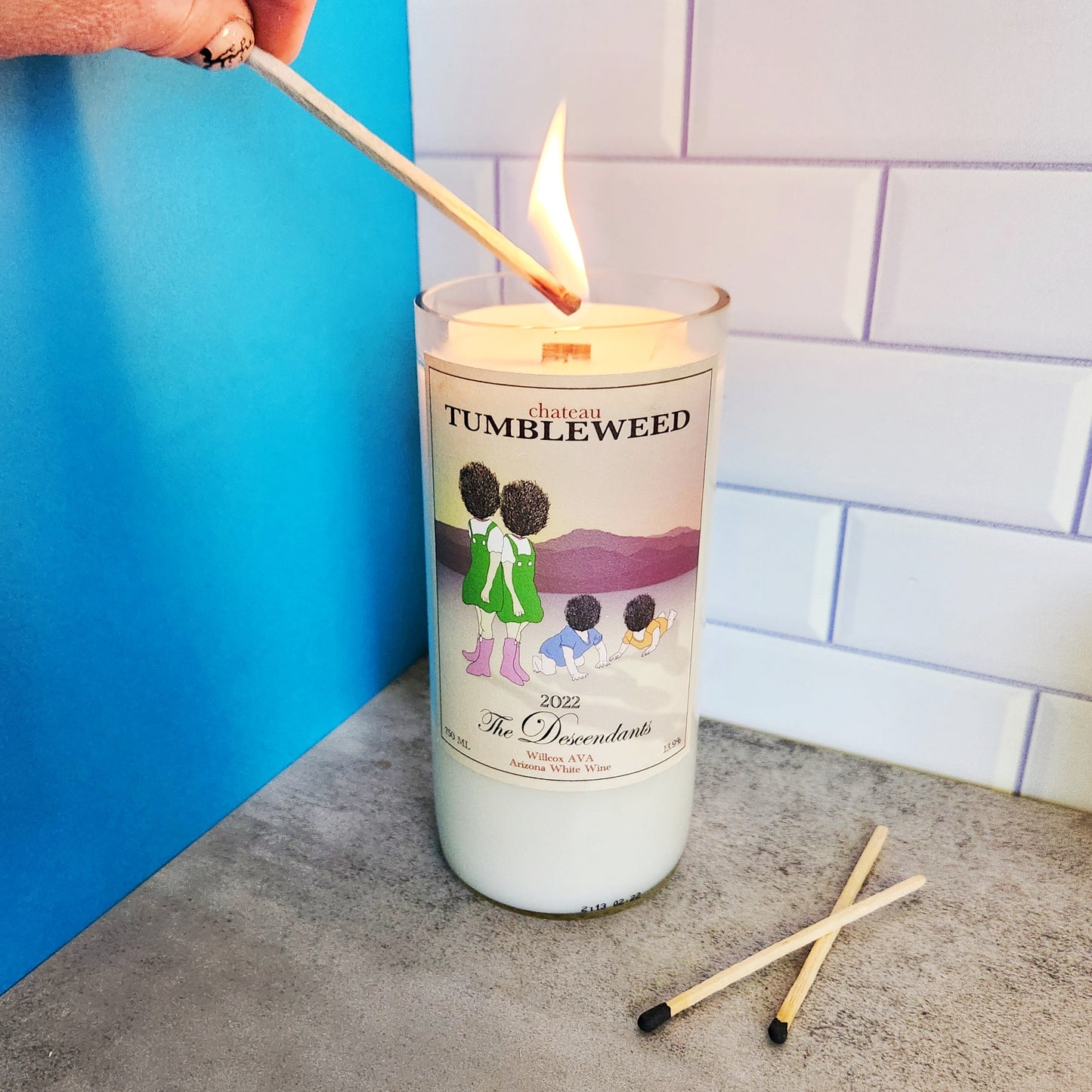 Hand lighting a Cactus Vine wine bottle candle in a recycled Arizona wine bottle with a Chateau Tumbleweed label. The candle is positioned on a light gray surface with two matches nearby, and the background features blue and white tile elements.