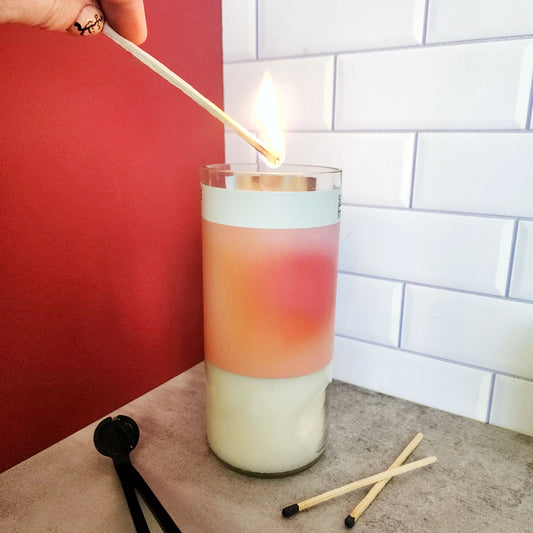 Hand lighting a Cactus Vine wine bottle candle in a recycled Arizona wine bottle with a frosted and gradient color label. The candle is set on a light gray surface with two matches and a wick trimmer nearby, and the background features red and white tile elements.