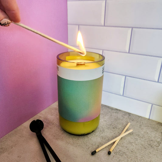 Hand lighting a Cactus Vine wine bottle candle in a recycled Arizona wine bottle with a gradient pastel label. The candle is set on a light gray surface with two matches and a wick trimmer nearby, and the background features lavender and white tile elements.