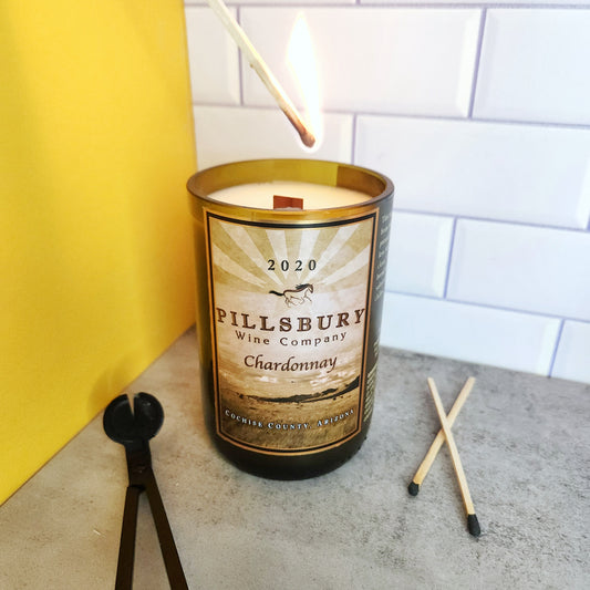 Hand lighting a Cactus Vine hand-poured, eco-friendly wine bottle candle in a recycled Pillsbury Wine Company bottle with a Chardonnay label. This coconut soy candle is set on a light gray surface with two matches and a wick trimmer nearby. The background includes yellow and white tile elements, highlighting the sustainable and artisanal qualities of this wine bottle candle.