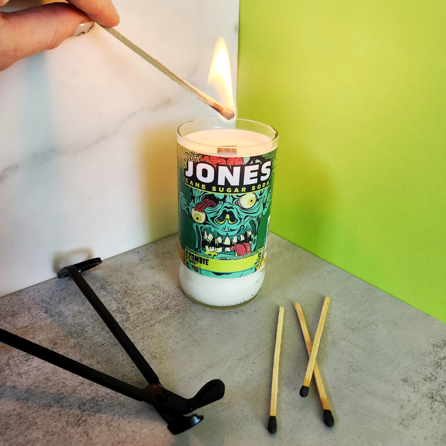 Lighting a Jones soda bottle candle with coconut-soy wax and wooden wick, showing eco-friendly Halloween decor with a zombie-themed label.