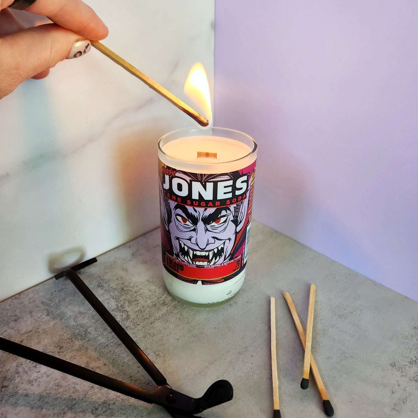 Lighting a recycled soda bottle candle with a vampire design, featuring coconut-soy wax and wooden wick for an eco-friendly Halloween vibe.