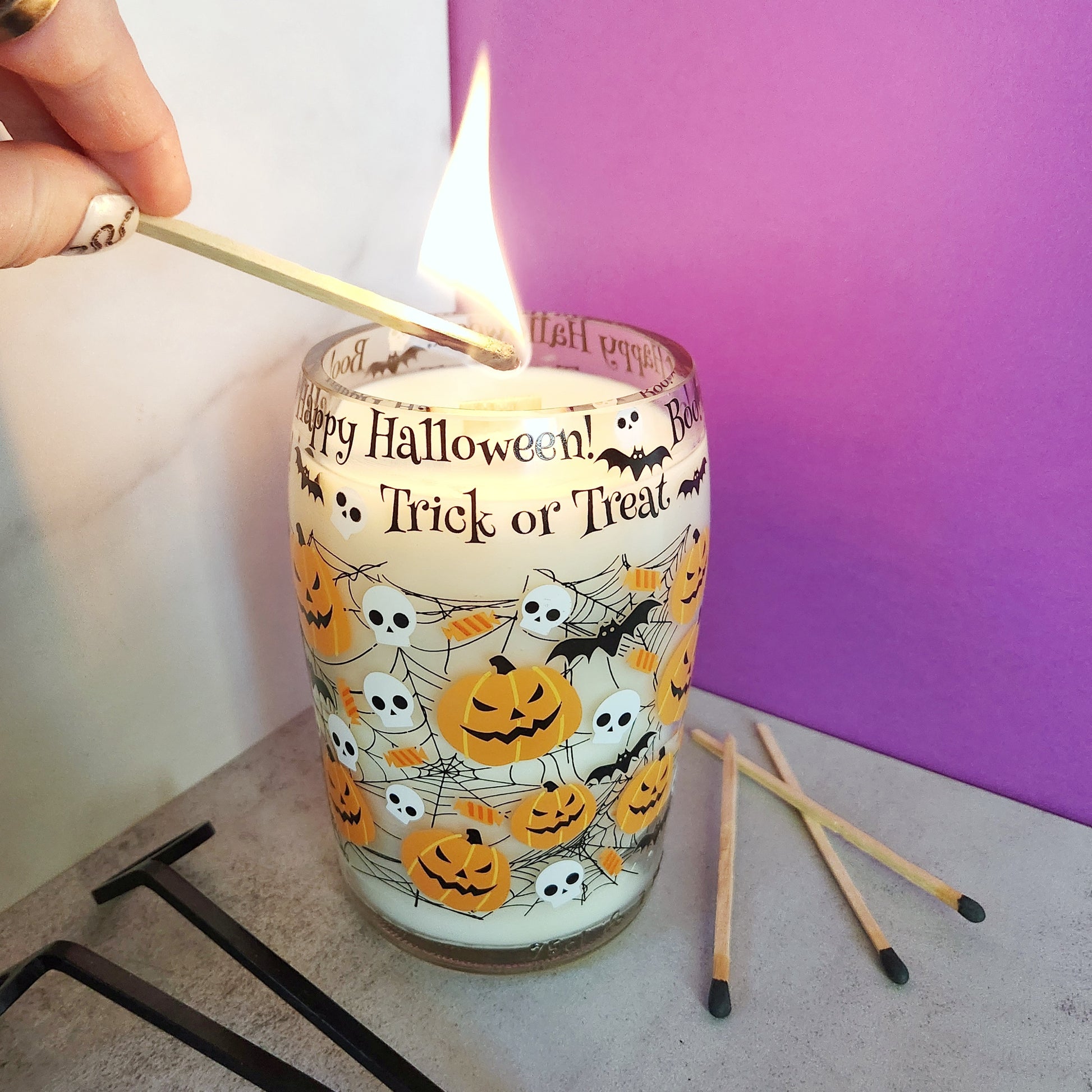 Lighting a Halloween-themed Caramel Apple scented wine bottle candle featuring jack-o'-lanterns, bats, and skulls with a match, showcasing the candle’s eco-friendly coconut soy wax and wooden wick.