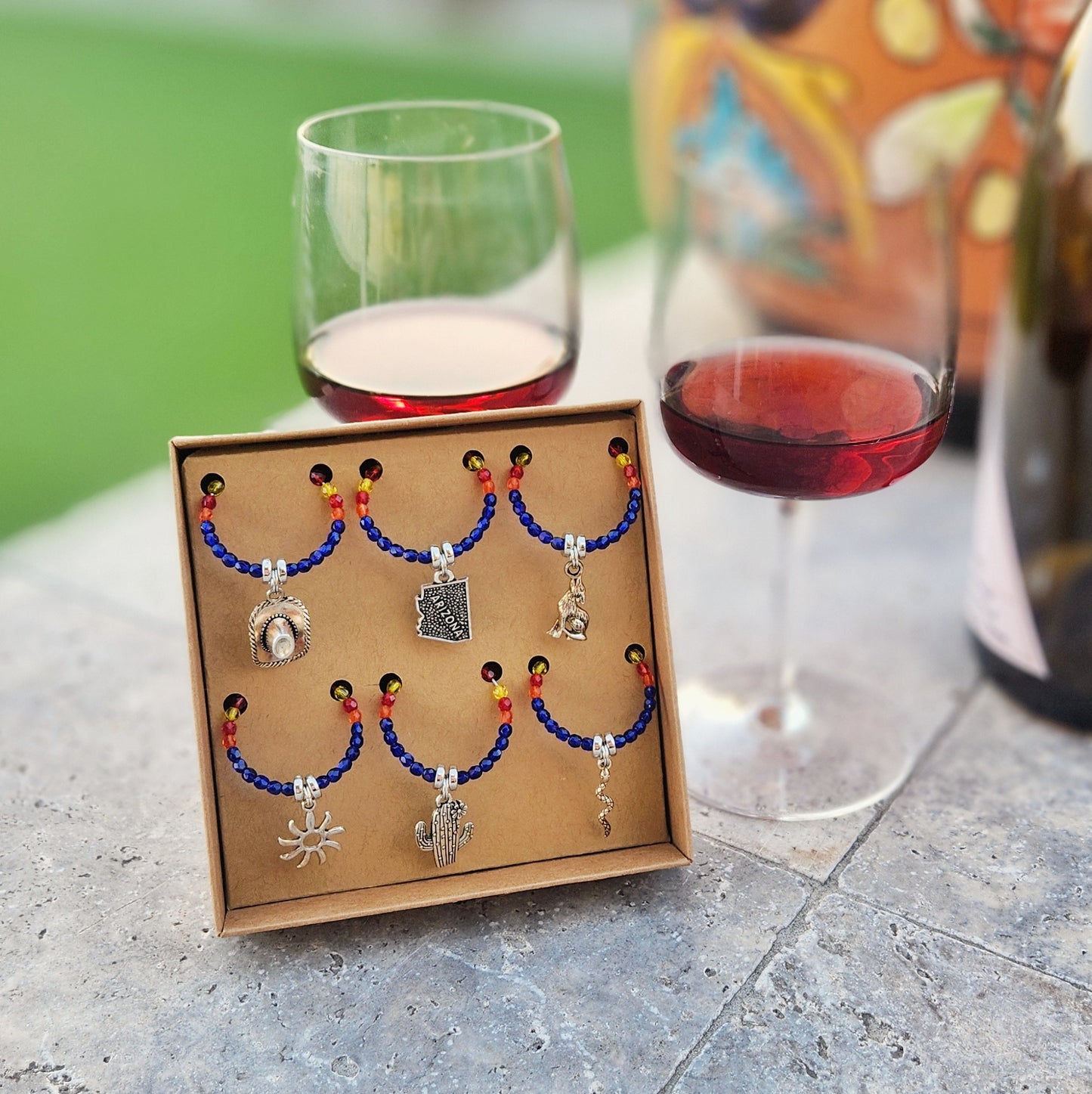 A set of colorful beaded wine glass charms with silver Arizona-themed pendants, including designs such as a cactus, snake, and cowboy hat, displayed in a cardboard gift box. The charms are placed on a stone surface alongside two glasses of red wine and a wine bottle, with a blurred green outdoor background adding a relaxed and inviting atmosphere.