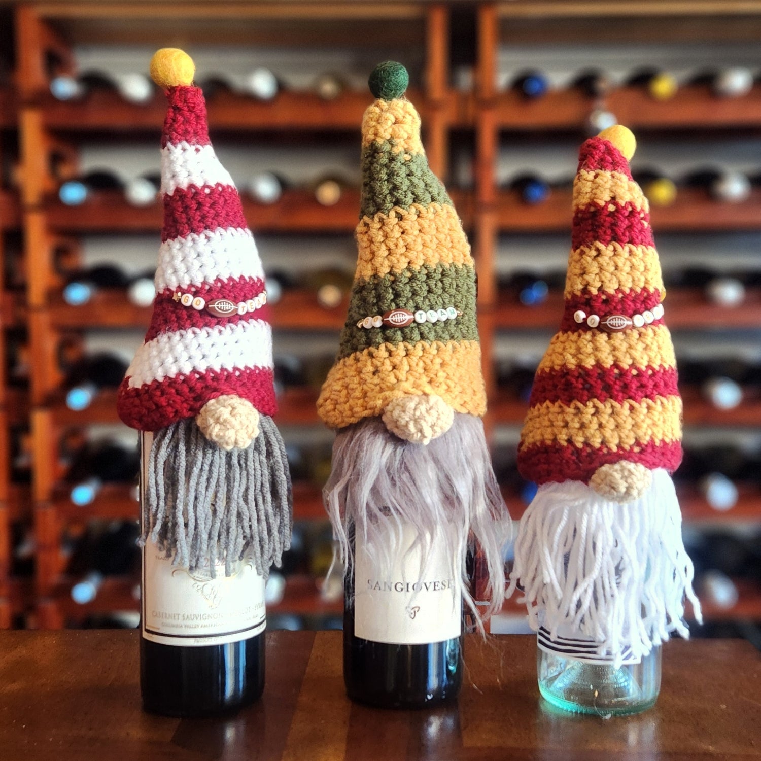 Three wine bottles with sports team theme crocheted gnome hat toppers. Each gnome  has beaded decorations, and long yarn beards in gray, white, and off-white add a whimsical touch. The backdrop includes a wooden wine rack.