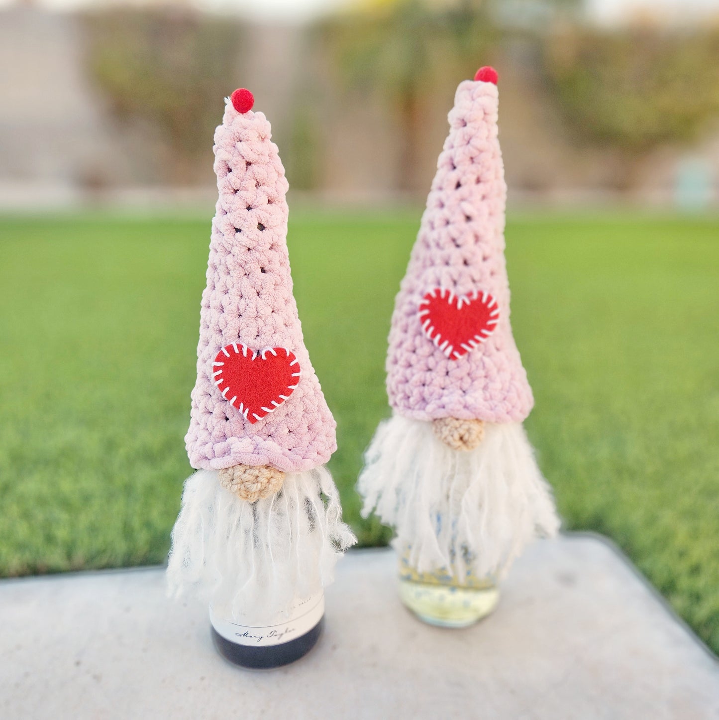 Two handmade Valentine-themed gnome bottle toppers displayed outdoors. Each gnome features a pink crocheted hat with a red felt heart stitched with white thread and a red pom-pom at the tip, symbolizing love and affection. They have long, fluffy white beards and rest on wine bottles.