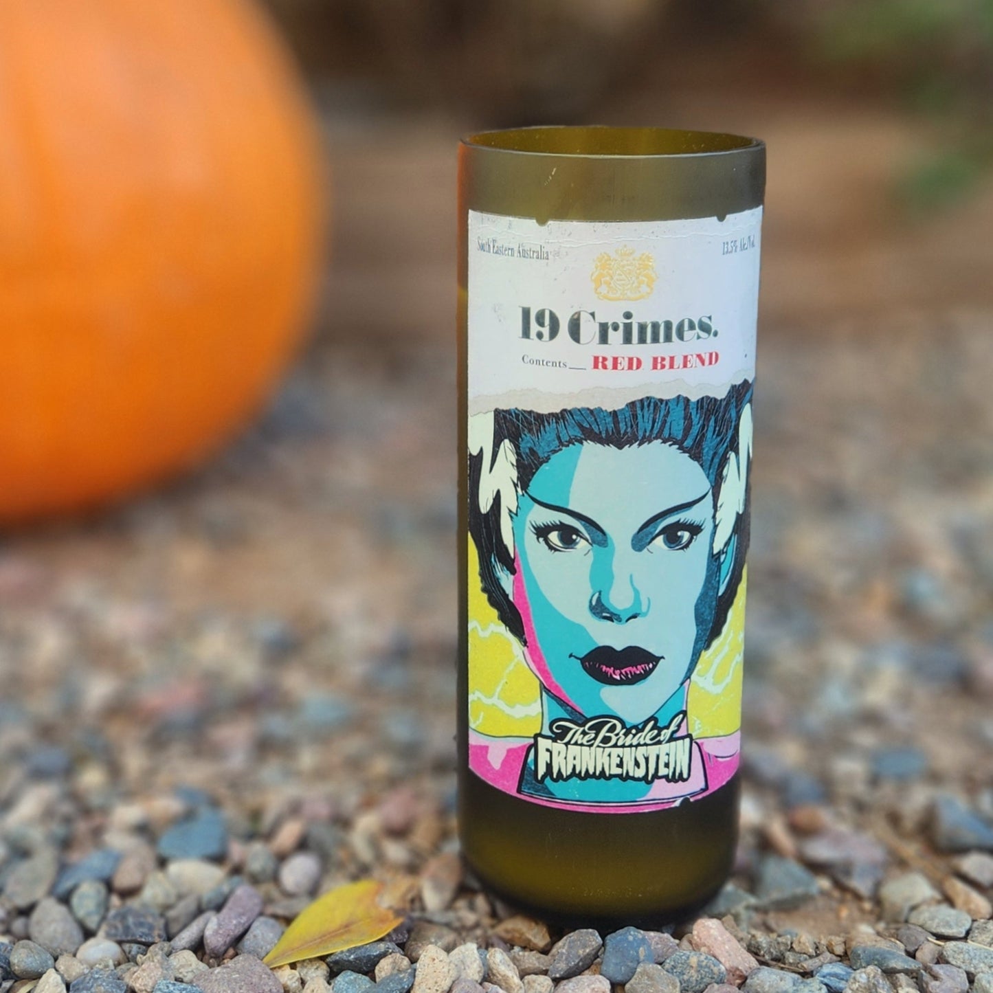 Close-up of a hand-poured candle in a recycled 19 Crimes red wine bottle, featuring vibrant pop-art style artwork of the Bride of Frankenstein. The eco-friendly candle sits on a gravel surface next to a pumpkin, highlighting its Halloween-ready vibe. The smooth, polished glass rim and visible wooden wick reflect the handmade craftsmanship and sustainable reuse of the wine bottle.