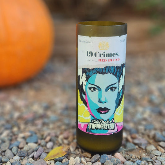 Close-up of a hand-poured candle in a recycled 19 Crimes red wine bottle, featuring vibrant pop-art style artwork of the Bride of Frankenstein. The eco-friendly candle sits on a gravel surface next to a pumpkin, highlighting its Halloween-ready vibe. The smooth, polished glass rim and visible wooden wick reflect the handmade craftsmanship and sustainable reuse of the wine bottle.