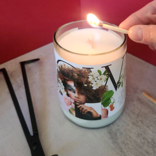 White candle made in a cut wine bottle.  Label features a magazine style cut out artwork of a woman with floral design.  Candle is being lit.