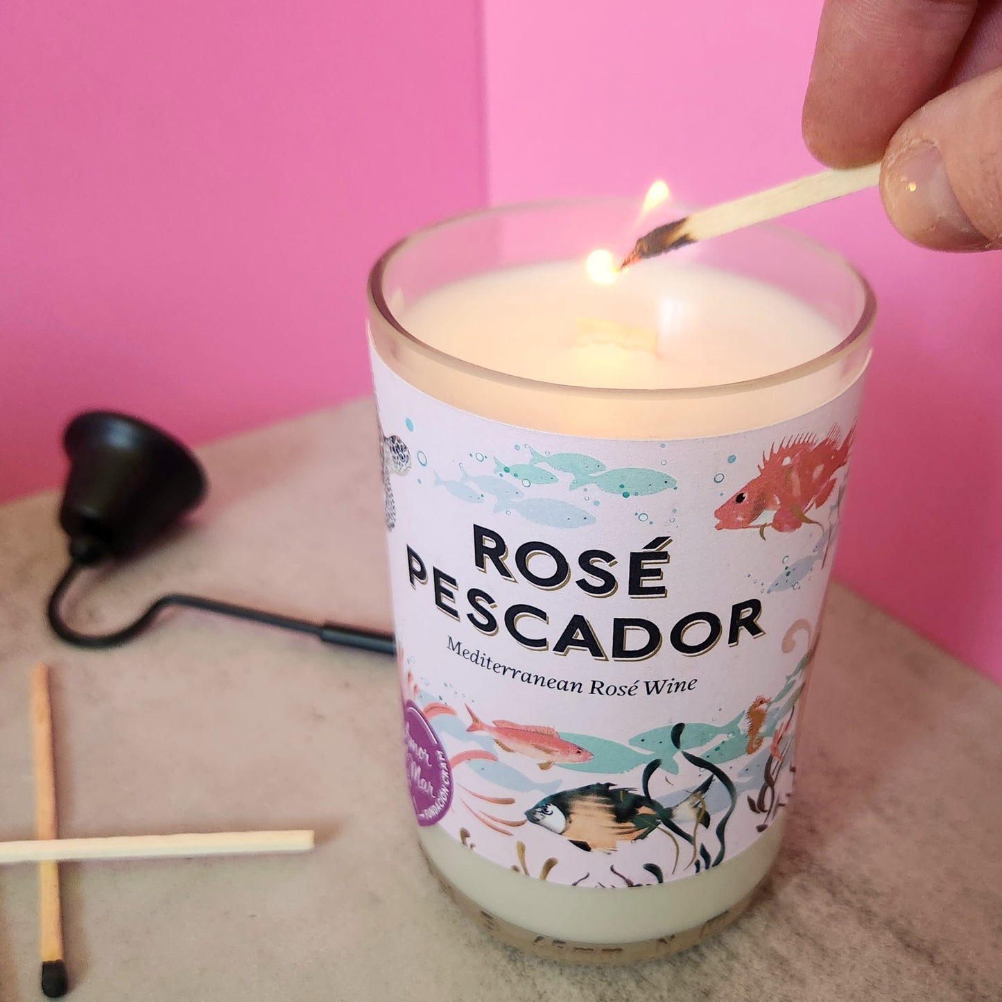 White candle made in a cut wine bottle.  Label stays Rose' Pescador Mediterranean Rose' Wine and has a pink background with drawings of sealife.  Candle is being lit.