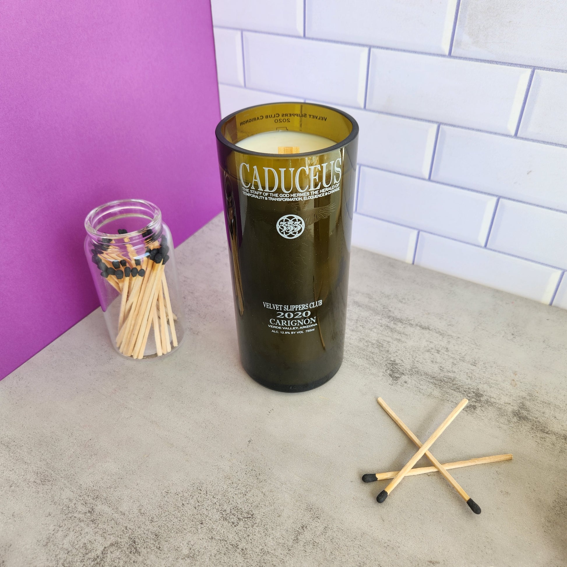 Display of the "Vanilla Twist" candle in a recycled wine bottle with matches and a purple background.