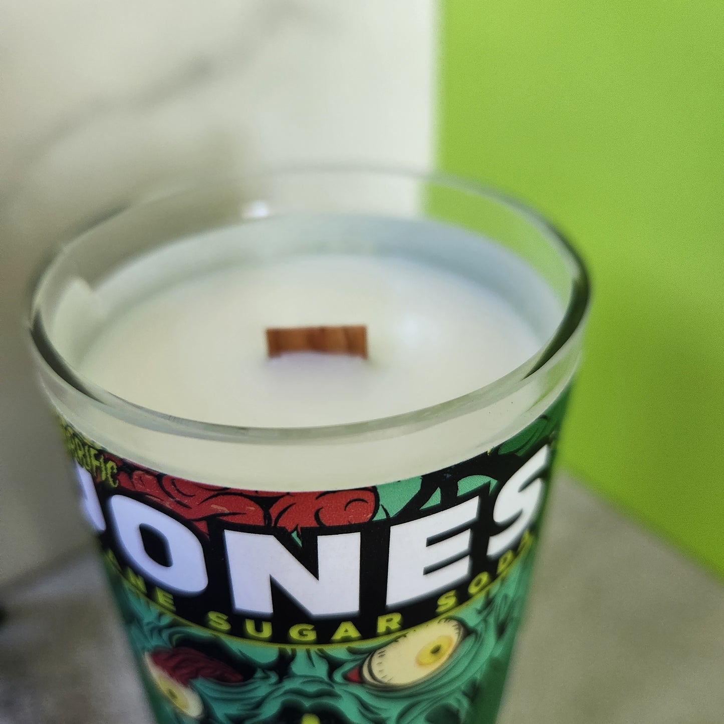 Close-up of wooden wick in hand-poured coconut-soy wax candle inside a recycled Jones soda bottle with vibrant zombie design for Halloween decor.