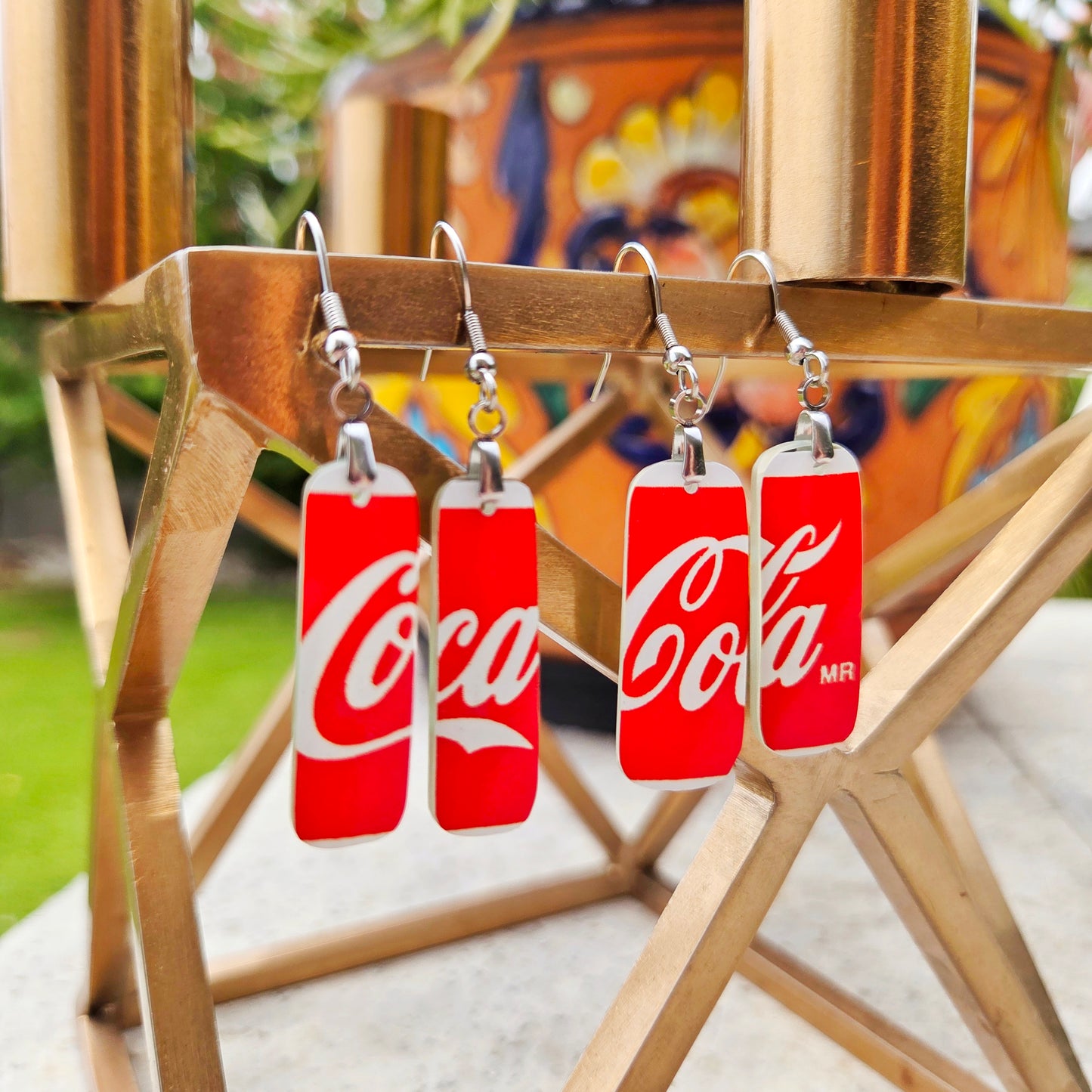 Four Coca-Cola themed drop earrings with red and white rectangular charms shaped like soda cans, hanging from silver fishhook wires on a wooden stand.