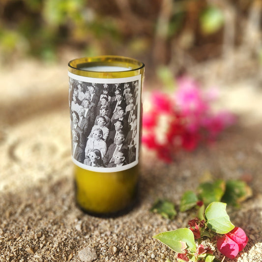 A recycled green wine bottle candle with a unique black-and-white vintage-style label depicting a group of people wearing 3d glasses. The candle sits on sandy ground, surrounded by scattered greenery and vibrant pink bougainvillea flowers in the background, creating an eclectic and artistic atmosphere.