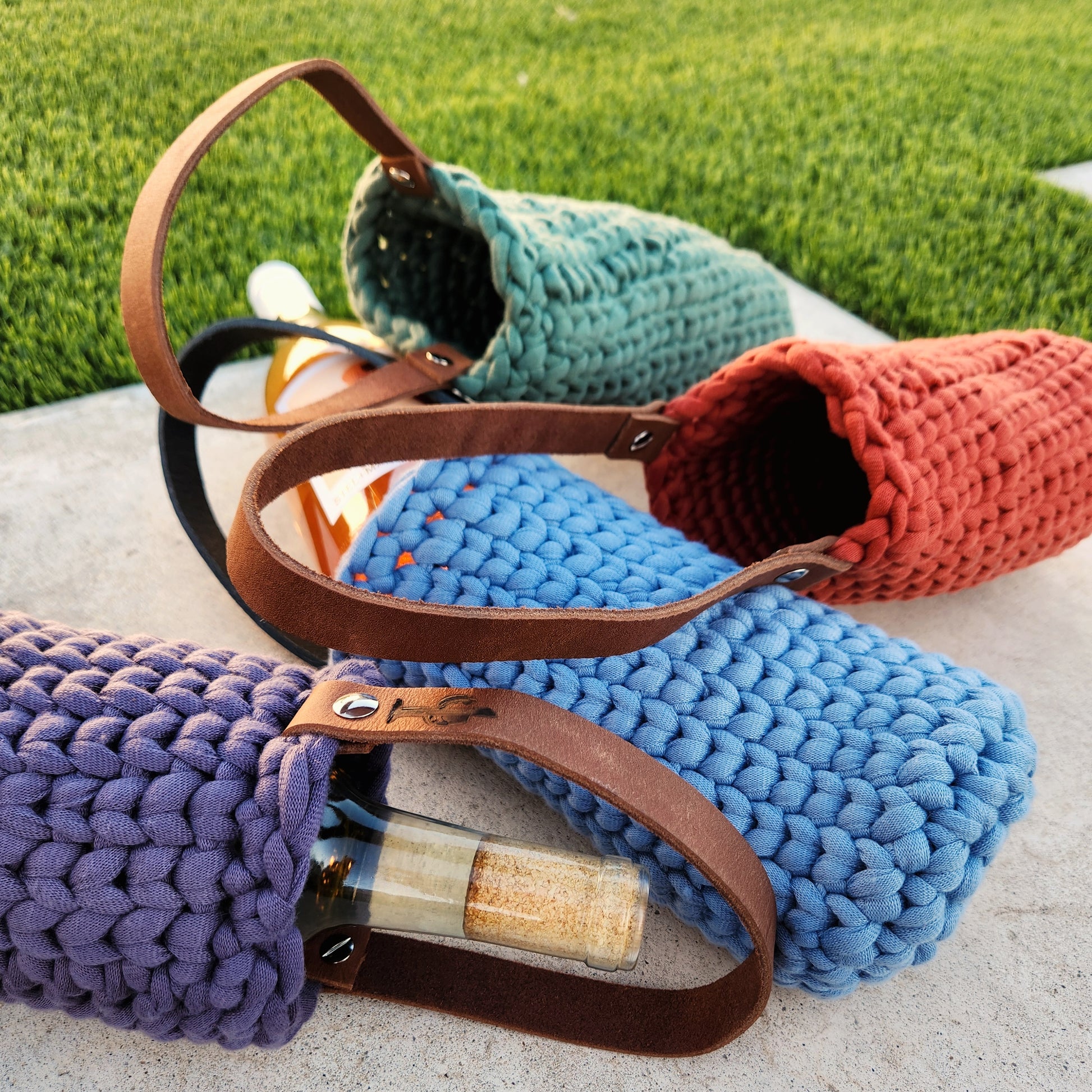 Four colorful crochet wine bottle carriers—purple, denim blue, sage green, and burnt orange—lay casually on a stone surface outdoors, each holding or beside a wine bottle. The hand-knit holders feature leather straps stamped with a cactus design, blending eco-conscious charm and desert-inspired style.