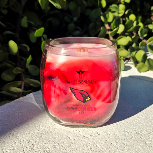 Milk Bottle candle with a red wax swirl and wooden wick.  Cardinals logo is on the front of the candle.  It's placed on a white surface with greenery behind it.