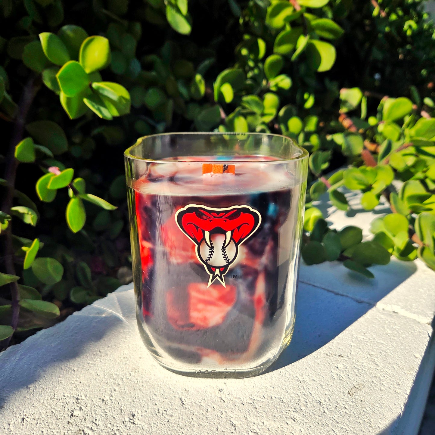 Milk bottle candle features red, black and teal swirled wax and a wooden wick.  Bottle features the AZ Diamondbacks logo and is set in front of greenery.