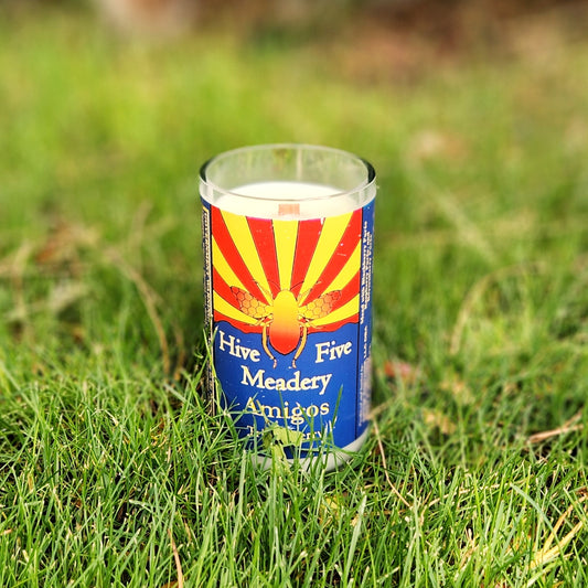 Hand-poured coconut soy wax candle in a recycled Hive Five Meadery bottle with bold Arizona state flag art, photographed outdoors on vibrant grass.