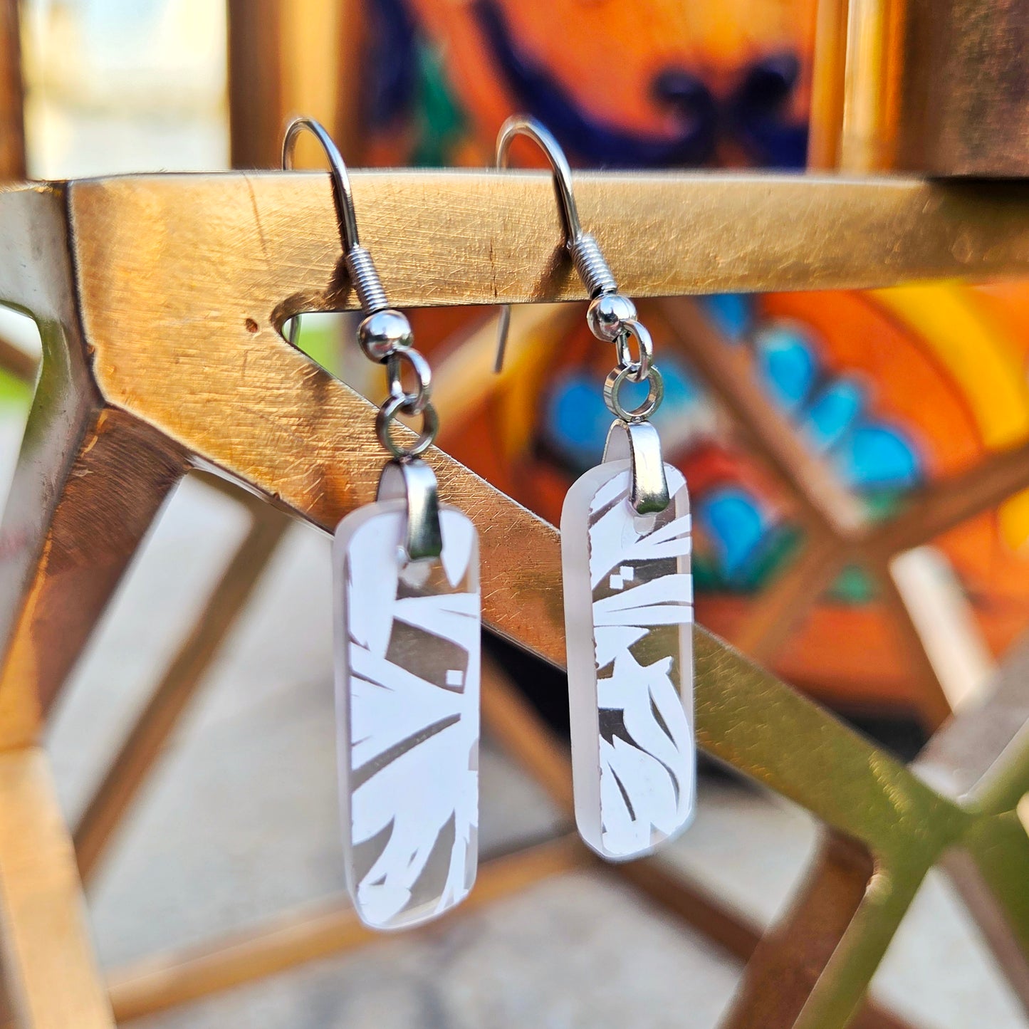 A pair of silver-toned drop earrings with rectangular white glass pendants featuring leaf-like patterns, hanging from a gold-colored metal stand.