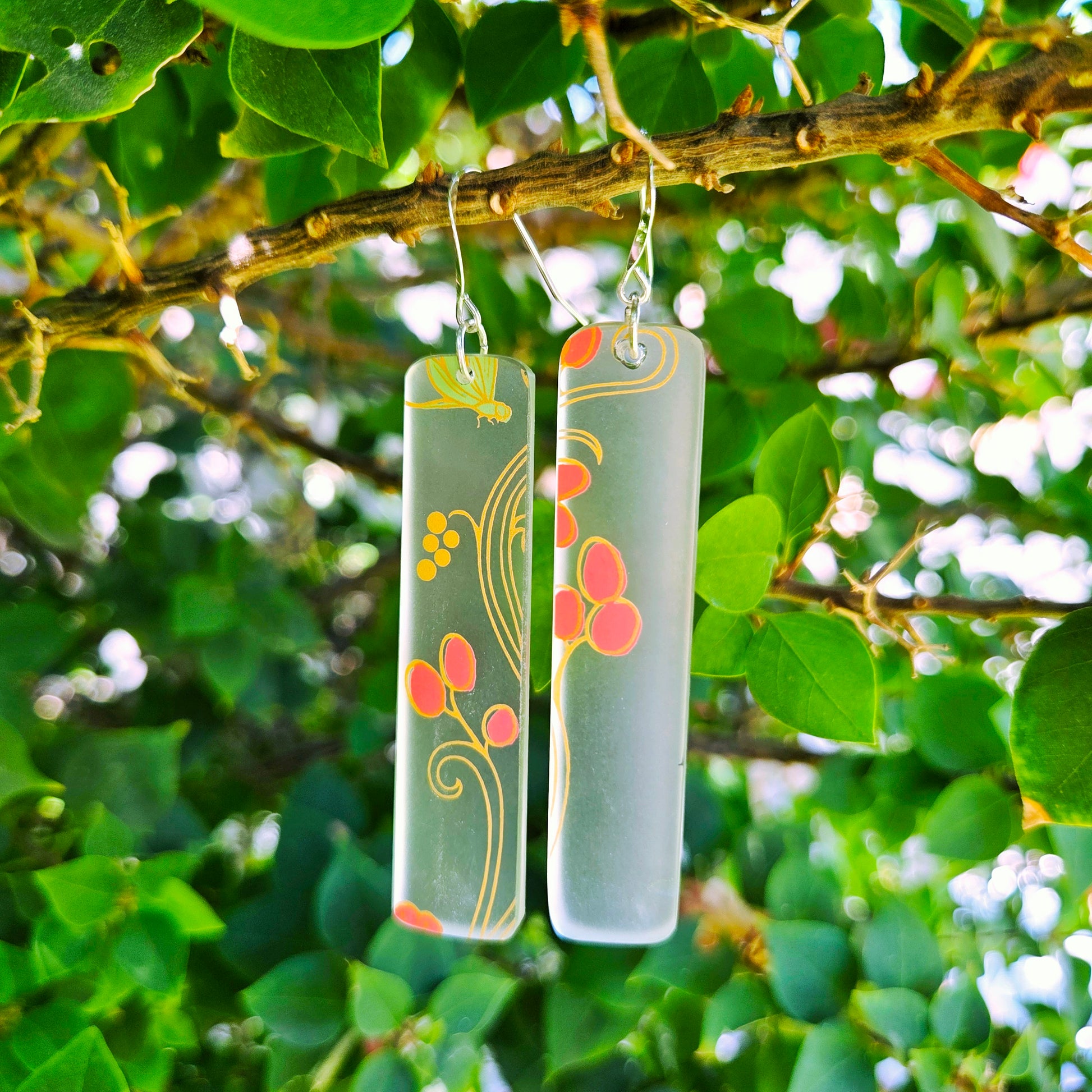 Frosted glass and coral vine earrings hanging from a branch in front of greenery.