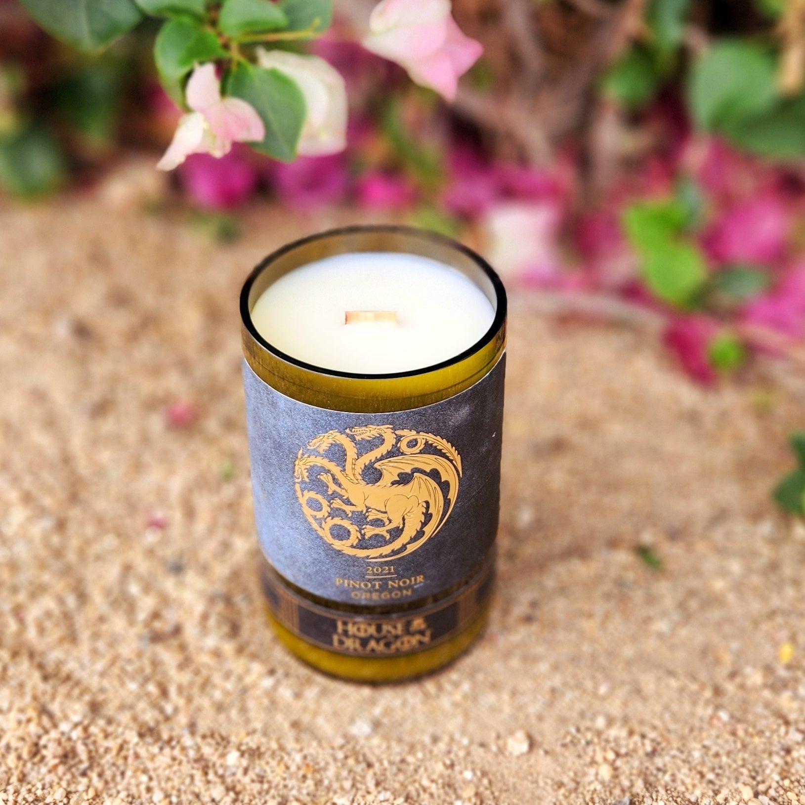 Angled top view of a hand-poured coconut soy wax candle in a recycled wine bottle with a wooden wick, featuring a House of the Dragon-style Pinot Noir label and a glossy glass rim, set against desert sand and pink bougainvillea.