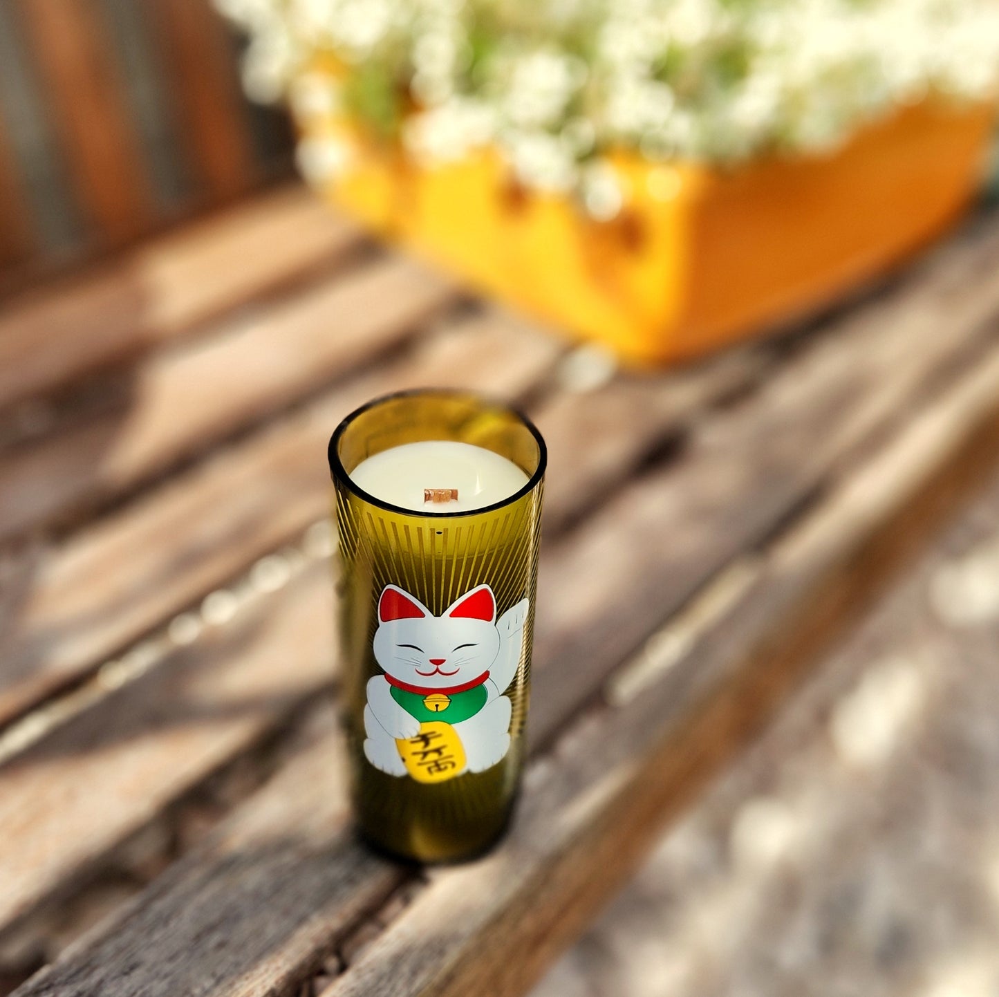 Eco-friendly wine bottle candle with a lucky cat design, made from a recycled green glass bottle and hand-poured with coconut soy wax. Features a clean-burning wooden wick and a smooth, polished rim, styled outdoors on a wooden bench for a cozy, sustainable vibe.

