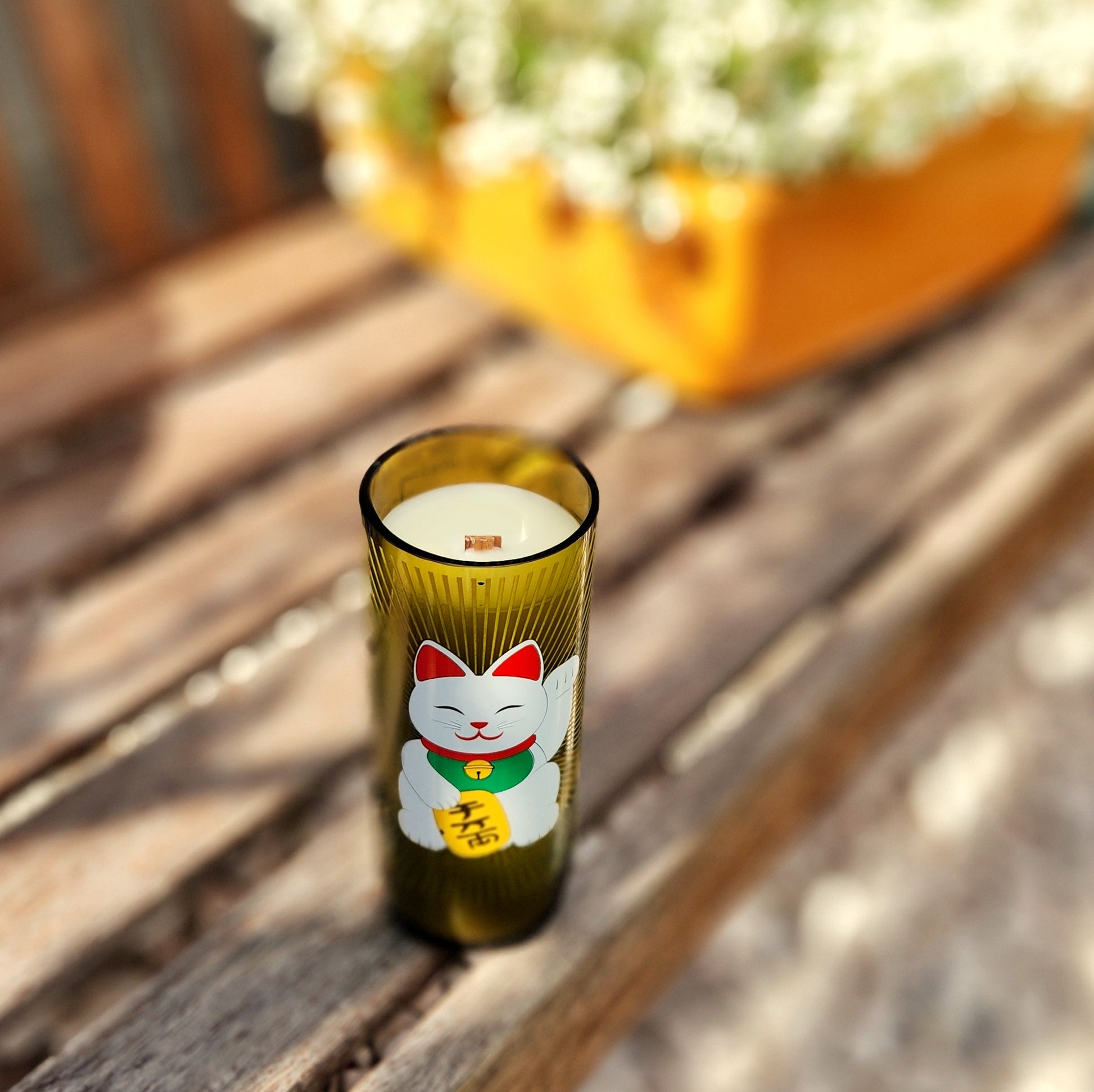 Eco-friendly wine bottle candle with a lucky cat design, made from a recycled green glass bottle and hand-poured with coconut soy wax. Features a clean-burning wooden wick and a smooth, polished rim, styled outdoors on a wooden bench for a cozy, sustainable vibe.


