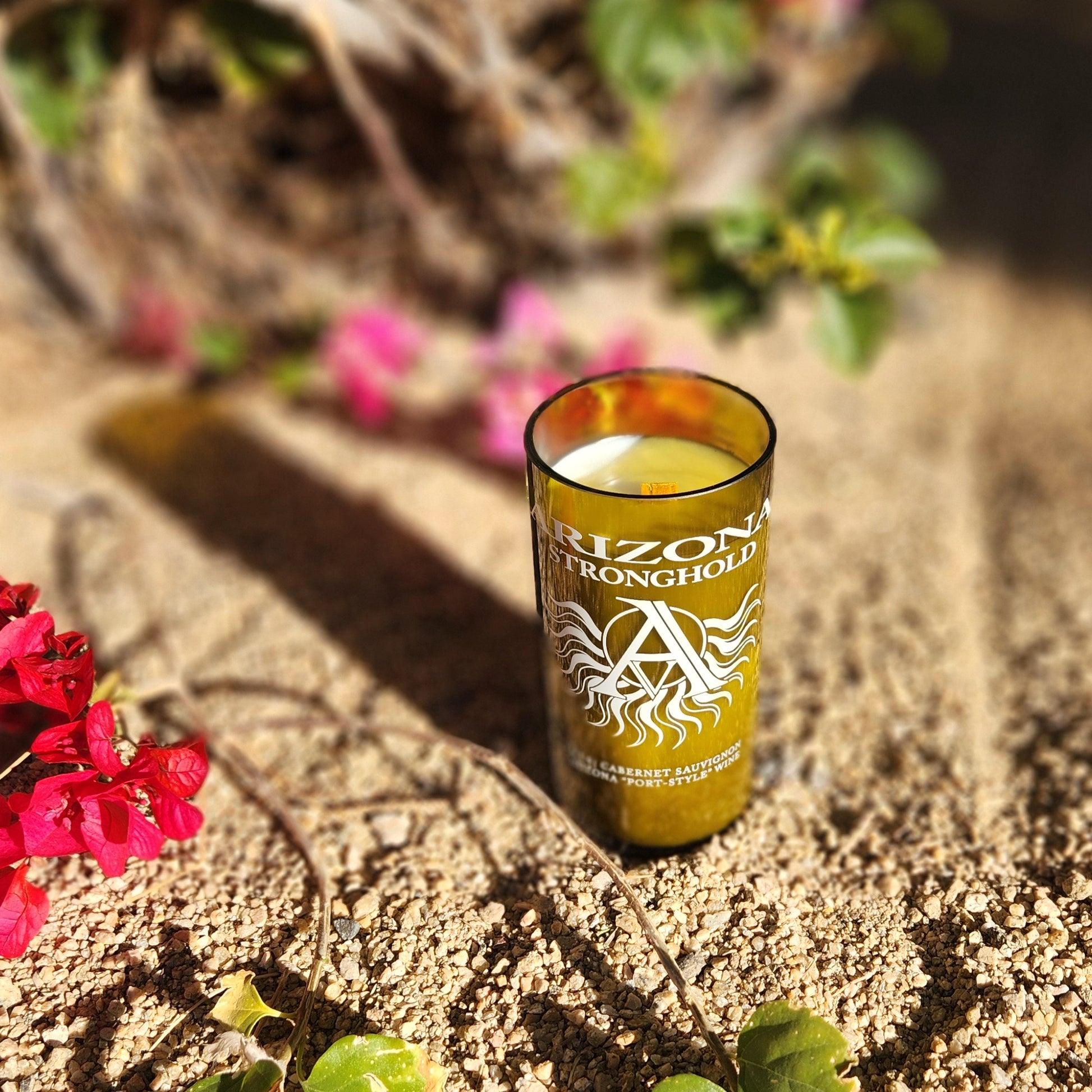 Angled outdoor view of a recycled Arizona Stronghold wine bottle candle resting on sandy ground with bougainvillea. The label’s bold white design and bright green glass reflect the candle’s artisan roots.