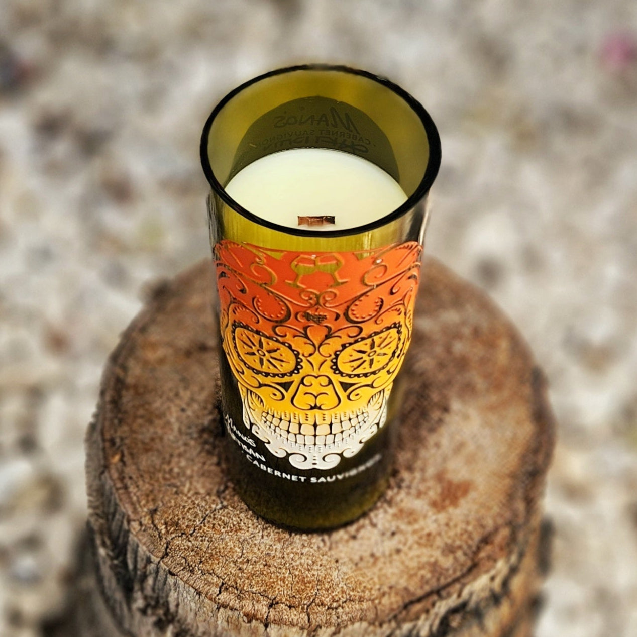 Angled overhead view of an eco-friendly coconut soy candle with a wooden wick housed in a recycled wine bottle. The front label features a vibrant sugar skull design, making this wine bottle candle a bold and sustainable decor piece.