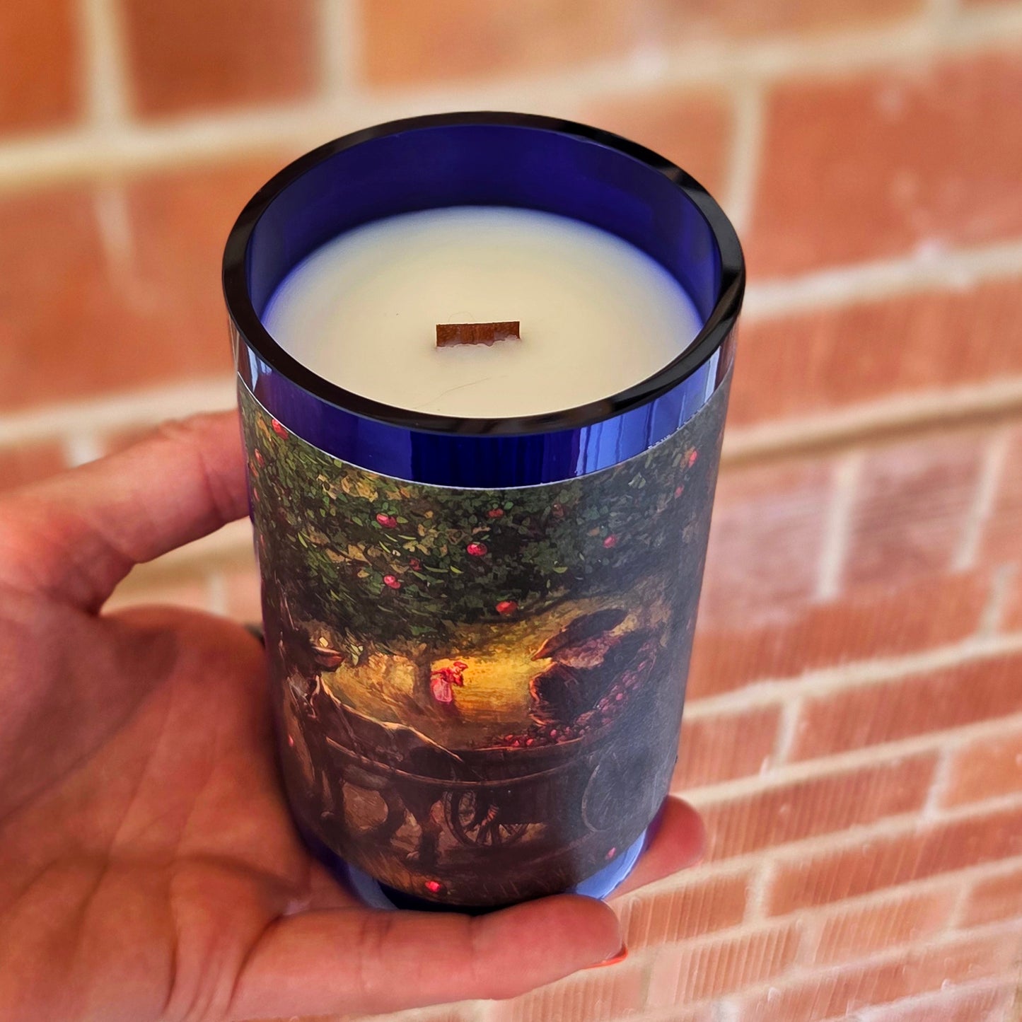 Angled shot of a blue glass wine bottle candle featuring orchard artwork, creamy white coconut soy wax, and a wooden wick, blending sustainability with vintage-inspired design.